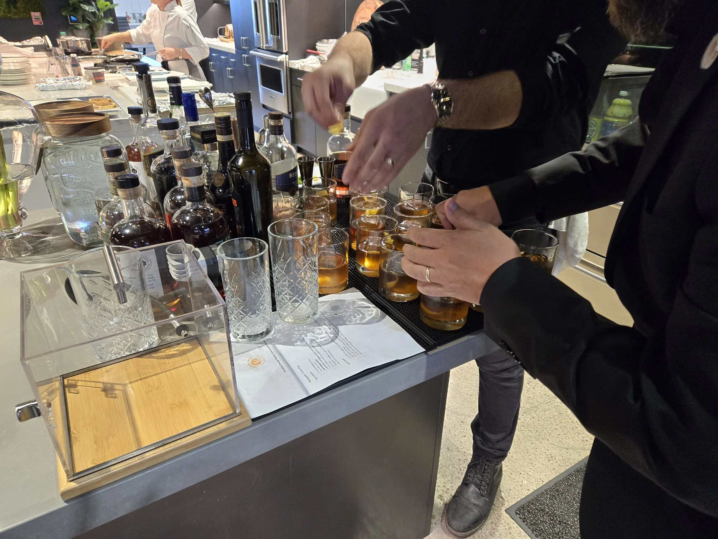 People preparing drinks at a bar or kitchen counter with bottles of alcohol and glasses filled with a dark amber liquid.