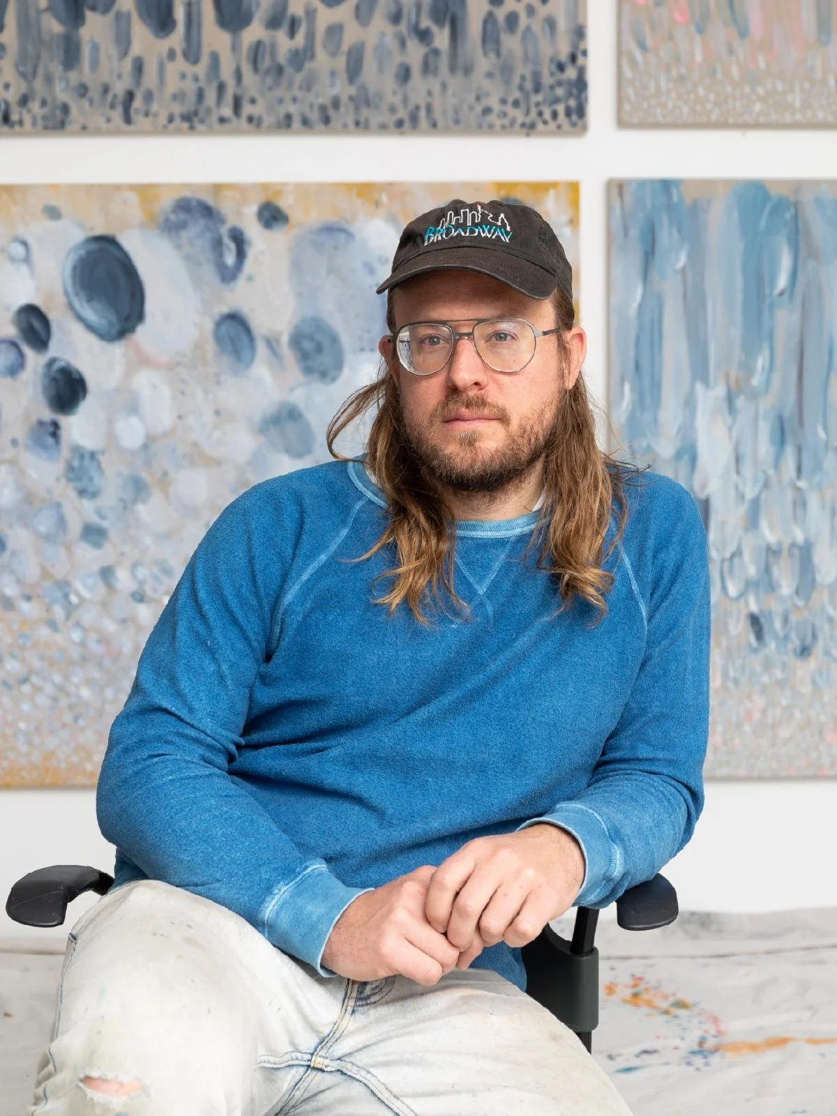 John Riepenhoff, a white man with long hair, glasses, and a beard, sits in a studio chair wearing a blue sweatshirt, paint-splattered pants, and a black baseball cap that reads “Broadway.” Behind him are several abstract, textural paintings in a soft