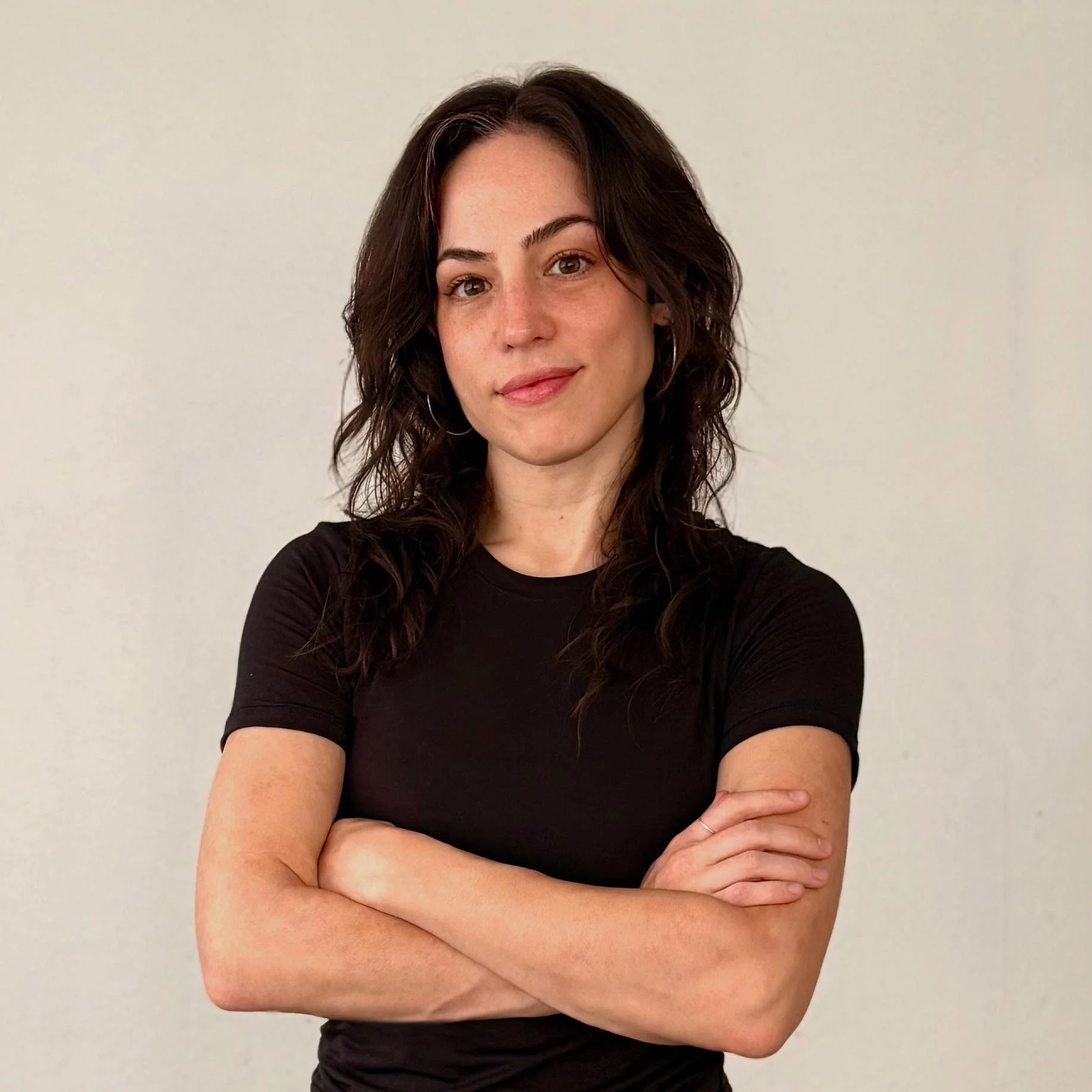 Rachel Shelton, a woman with long dark wavy hair, stands confidently with arms crossed in front of a plain light-colored background. She wears a fitted black t-shirt and gazes calmly at the camera with a slight smile.
