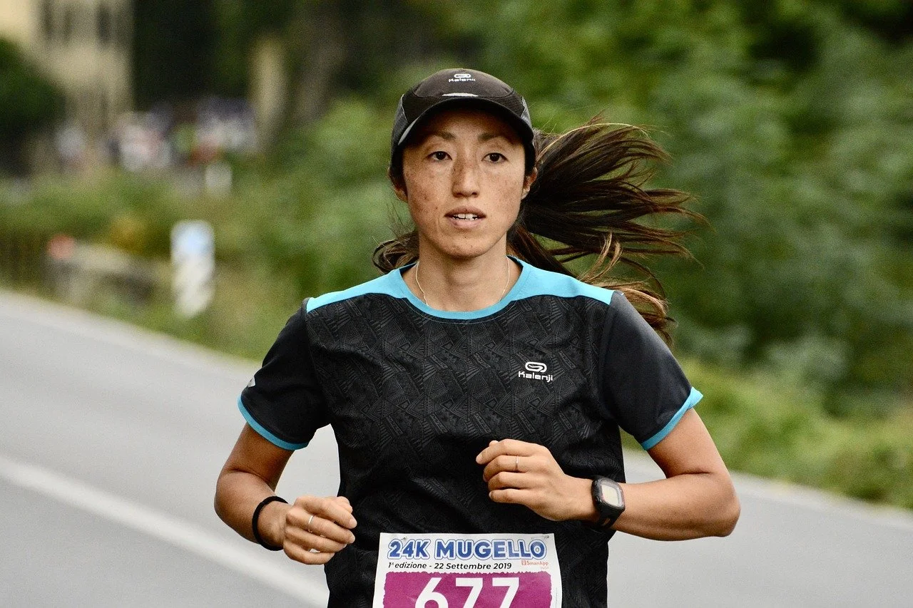 A woman running outdoors on a paved road during a race, wearing a black and blue athletic shirt, a black cap, and a wristwatch, with a race bib number 677 that reads '24K Mugello' and the date September 22, 2019.