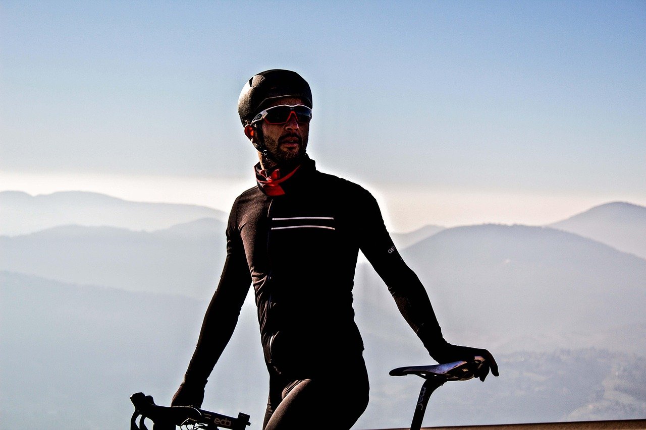 A man in black cycling gear, helmet, and sunglasses standing next to his bicycle with a mountain landscape in the background.