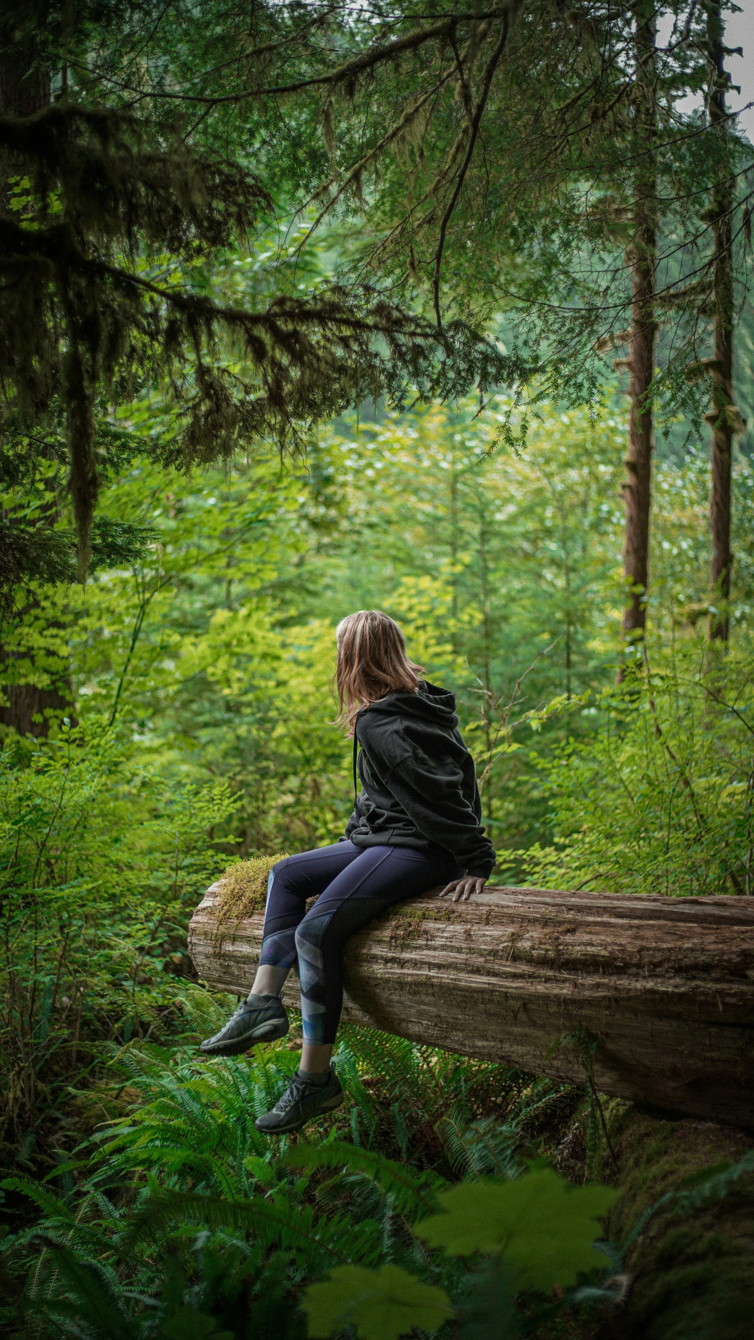 A woman wearing a black hoodie and dark leggings sitting on a fallen tree trunk in a lush green forest surrounded by dense trees and foliage.