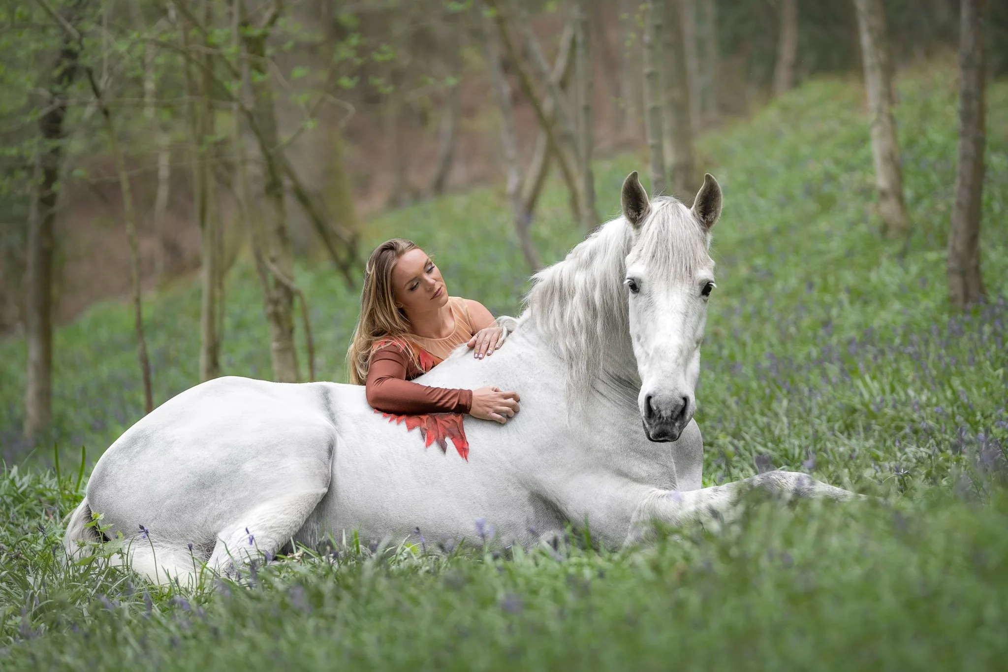 Portrait of horse stunt performer Georgia Plimbley embracing her white horse in a forest with bluebells