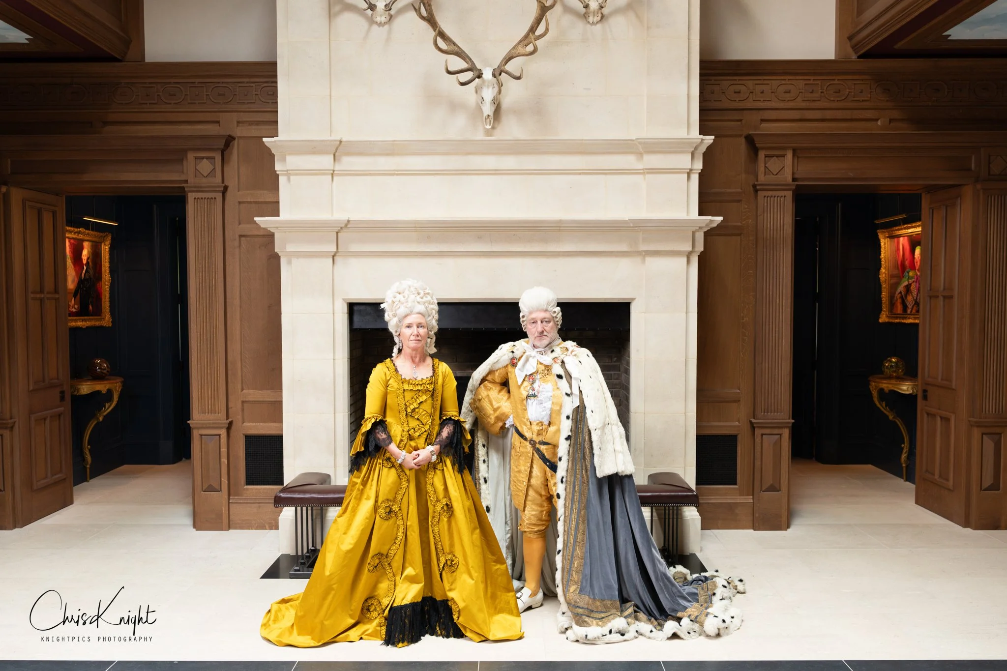 A man and woman dressed as Queen Charlotte and King George III standing in front of a grand fireplace in the entrance hall of an elegant mansion.