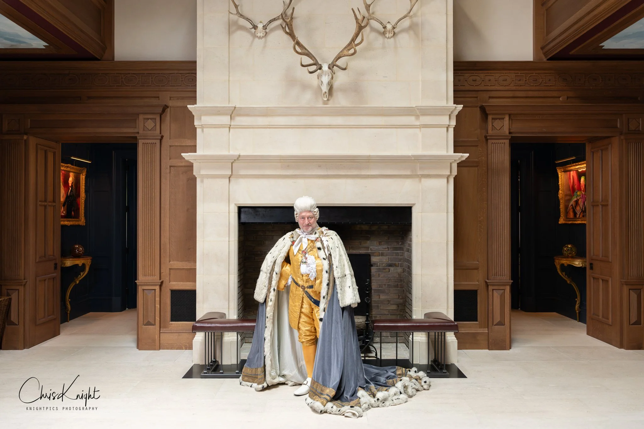 A man  dressed as King George III standing in front of a grand fireplace in the entrance hall of an elegant mansion.
