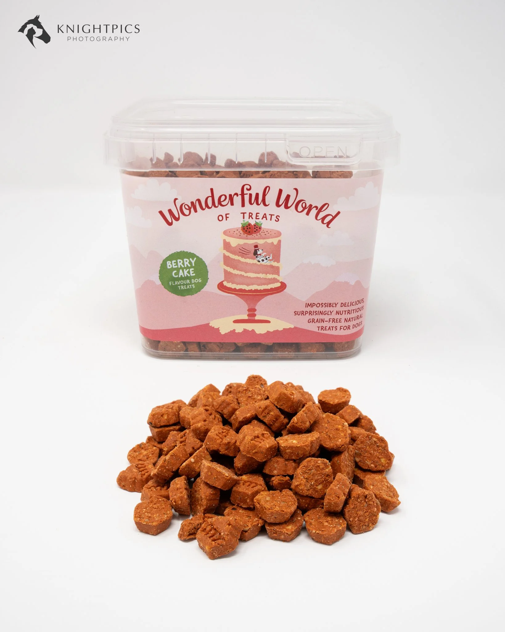 Professional commercial product photography featuring a pink tub of berry cake flavoured dog treats from The Wonderful World of Treats, with orange-coloured treats arranged in front. This clean studio photograph showcases the product's packaging, col