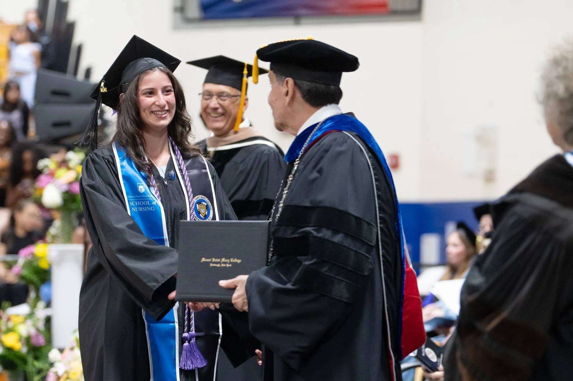 Graduate Julianna Zeidan shakes hands with President Dr. Robert Gervasi at Commencement. She is smiling wearing graduation regalia.