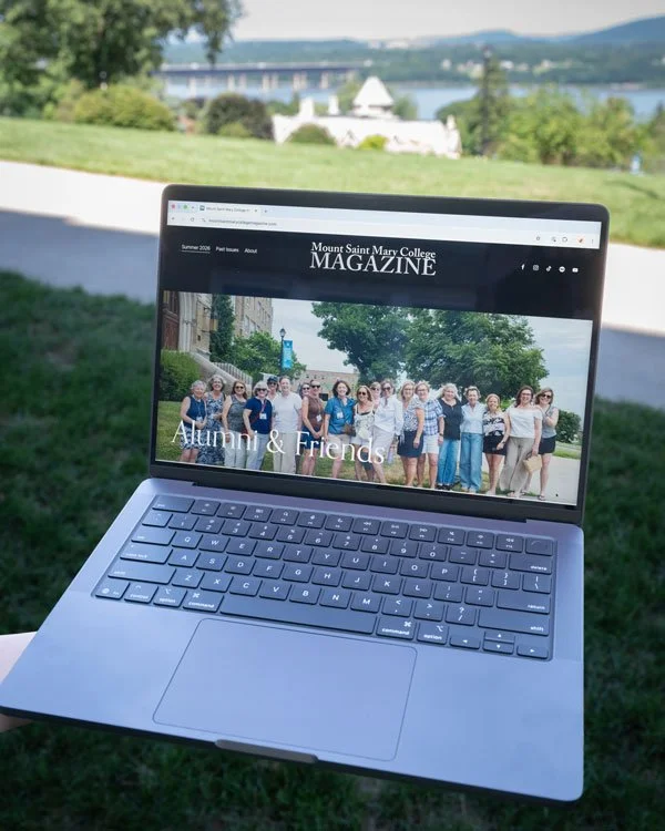 A laptop showing the homepage of Mount Saint Mary College Magazine with a photo of Mount alumni with the words "Alumni & Friends" across it. The laptop is outside with a view of the Hudson River and a bridge in the distance.