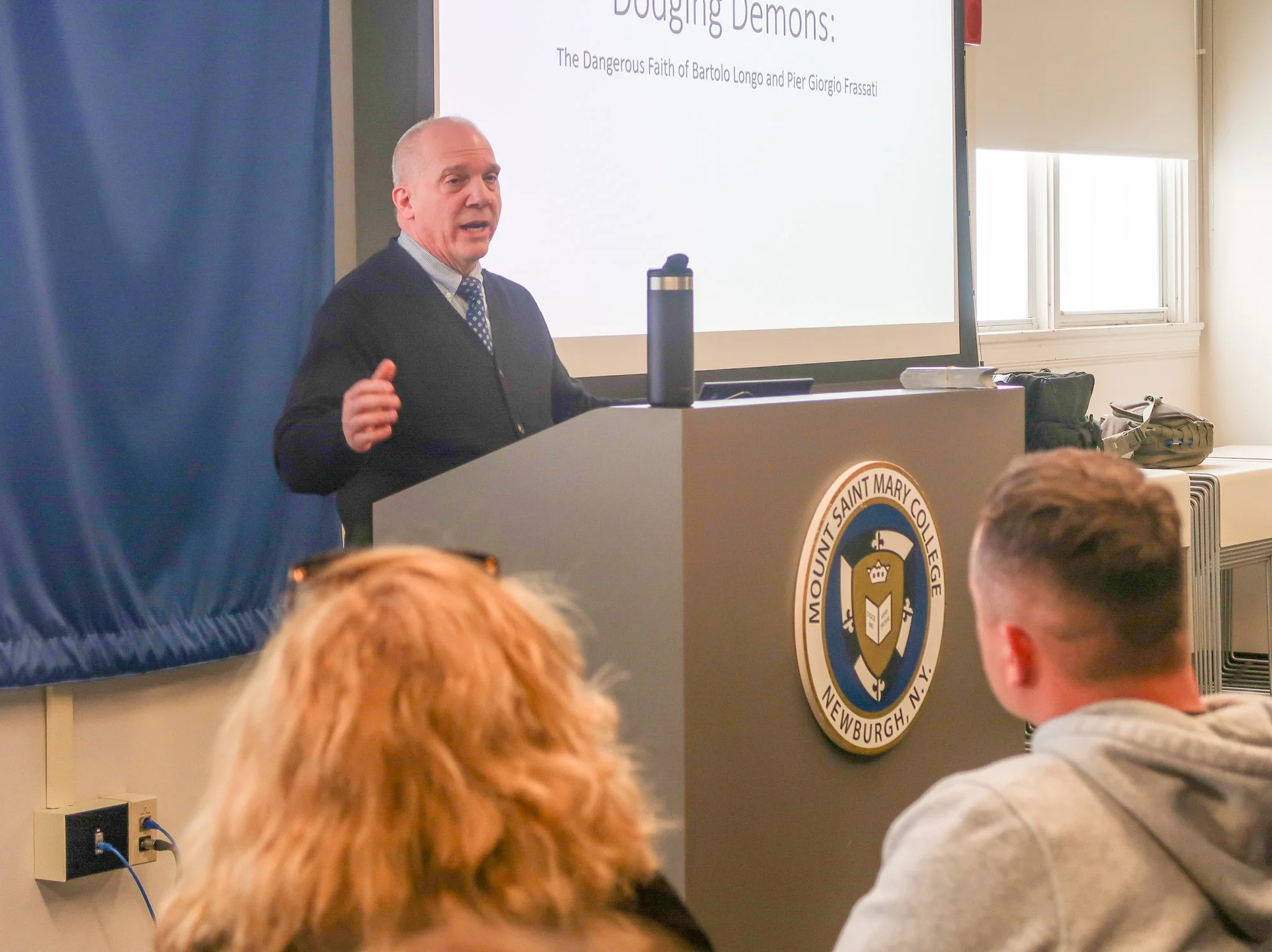 Robert Miller stands at a podium in the Dominican Center with his presentation on a slide behind him.