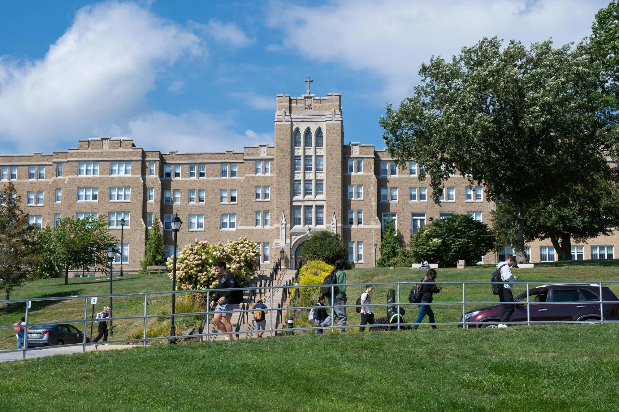 Students walk on the sidewalk between classes with the Dominican Center in the background on a sunny day.