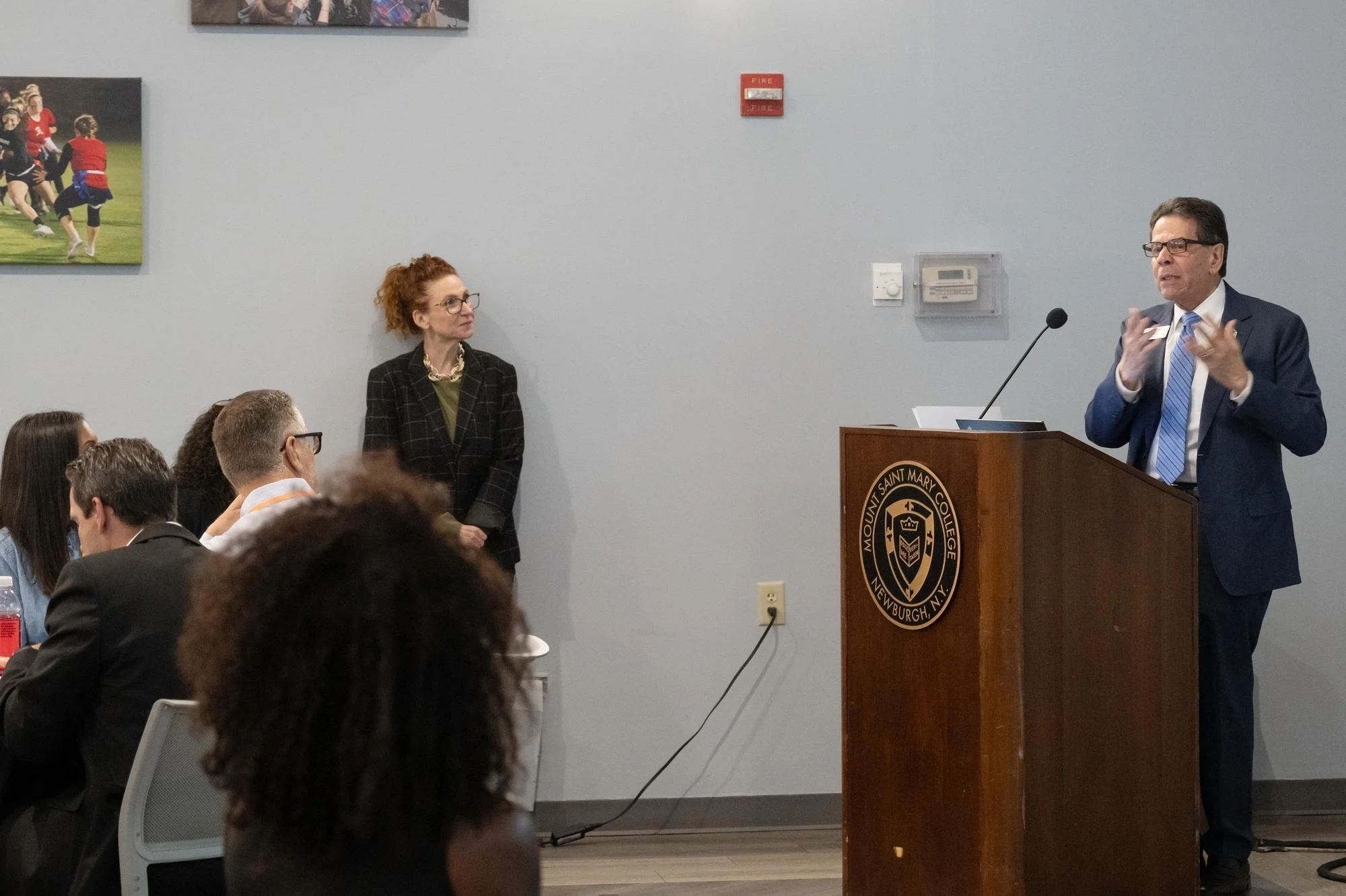   Mount President Dr. Robert Gervasi delivers his welcoming remarks at the lectern during the inaugural “Partners in Purpose” forum. Standing against the wall is Heather Bell, President and CEO of the Orange County Chamber of Commerce, who introduced