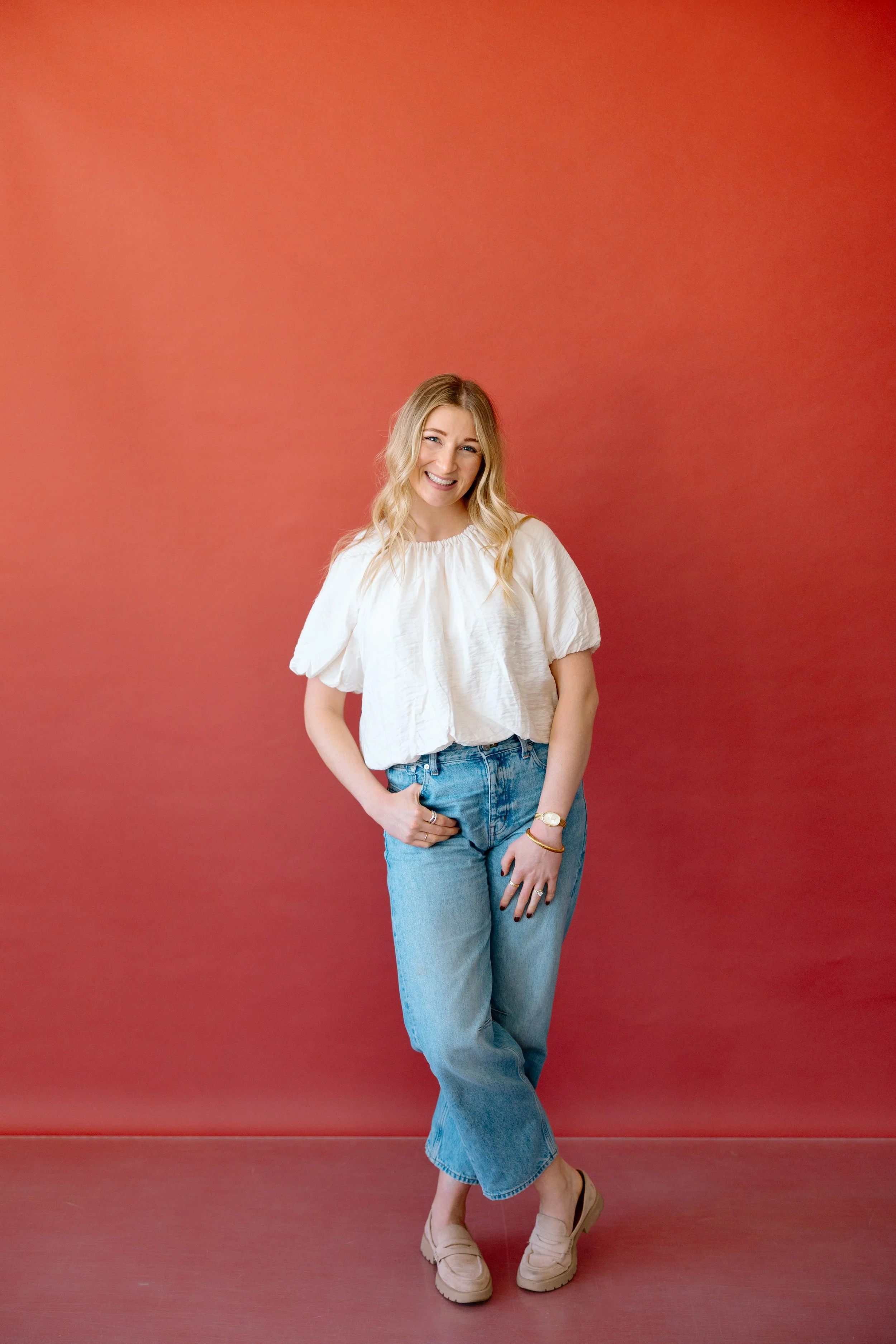 A woman with blonde hair wearing a white top, blue jeans, and beige shoes, standing and smiling against a solid red background.
