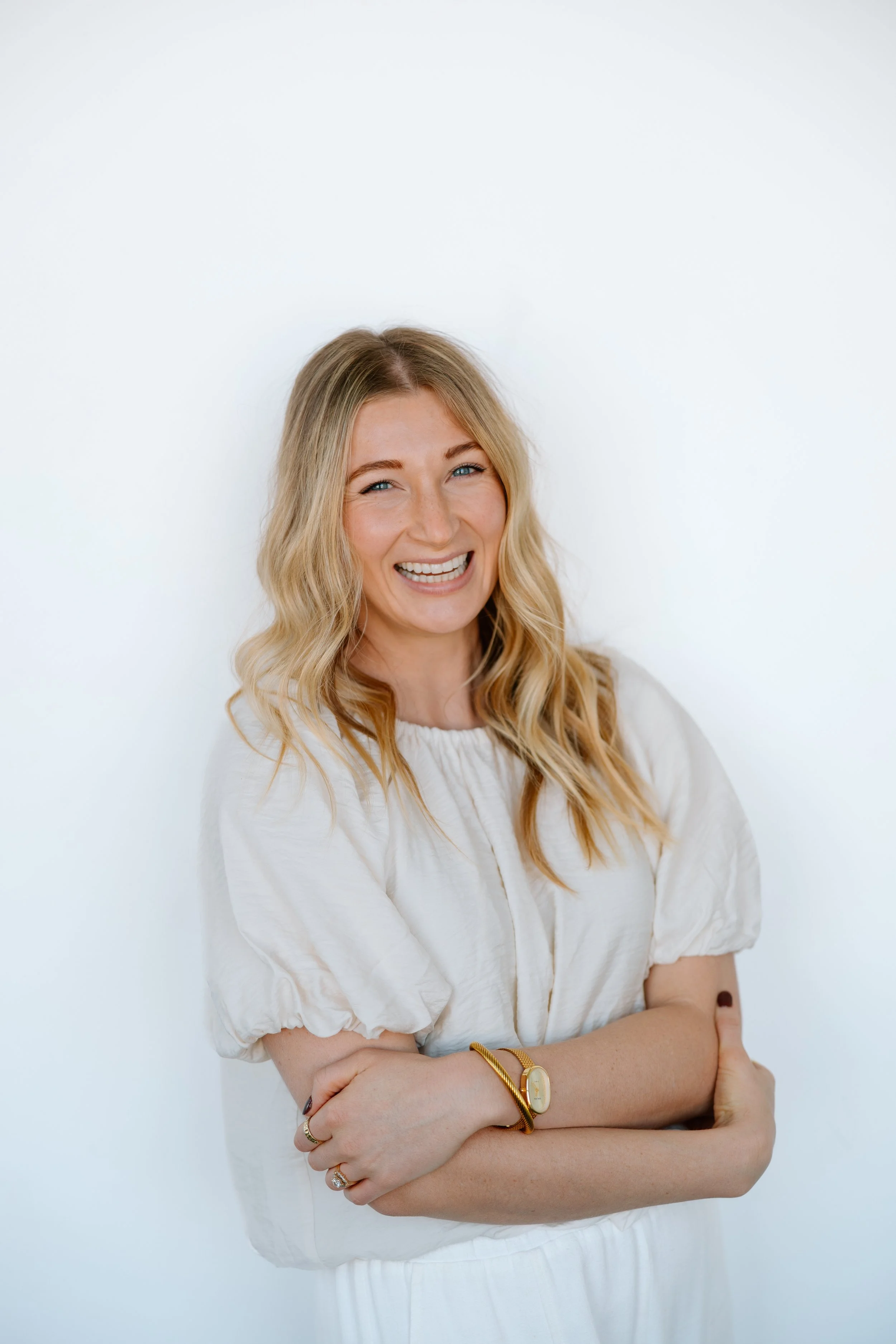 A smiling woman with long blonde hair, wearing a white blouse with puffed sleeves, standing against a plain white background.