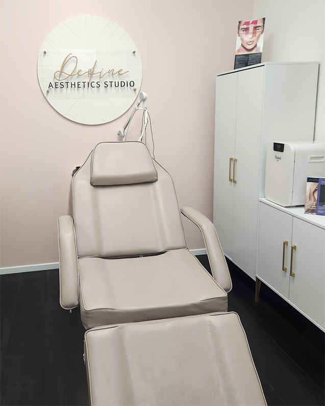 A beige medical or aesthetic treatment chair in a treatment room, with a headrest, in front of a wall with a sign that reads 'Define Aesthetics Studio'. There is a white cabinet with gold handles and a printer or sterilizer on top.