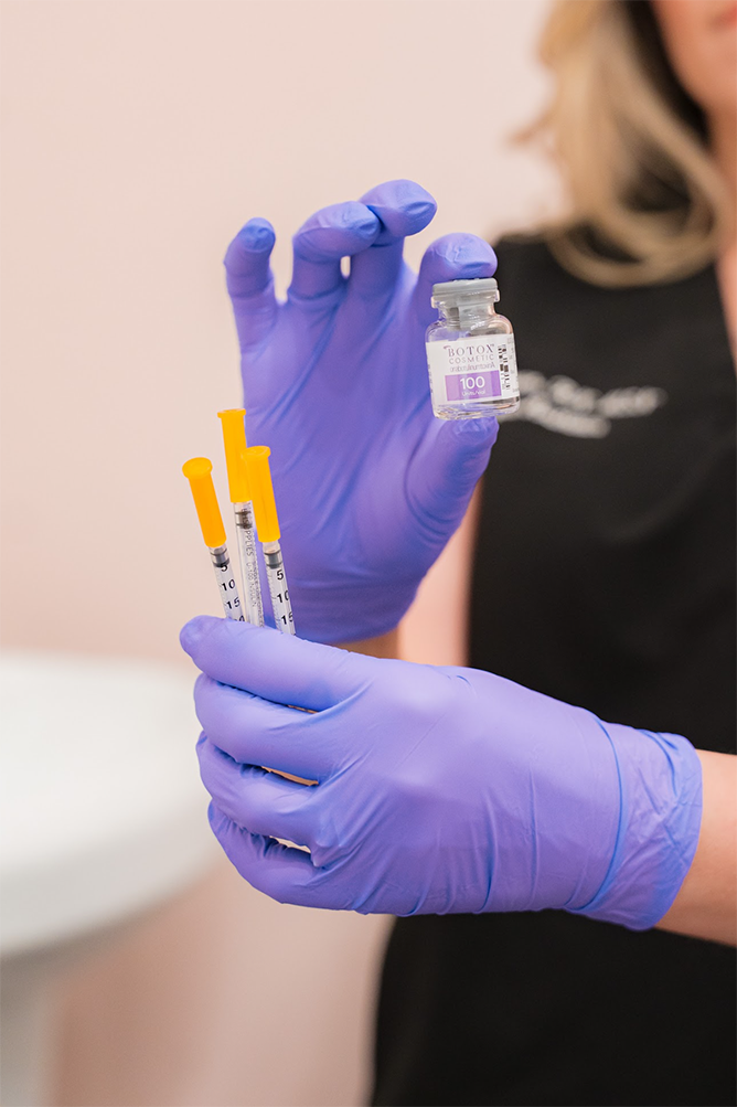 A person wearing purple gloves holding a small vial and three syringes with yellow plungers, likely in a medical setting.