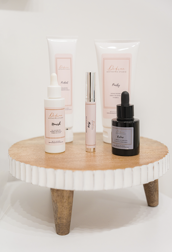 Skincare products on a small wooden table, including a white dropper bottle, a black dropper bottle, and a pink-capped tube, with blurred background