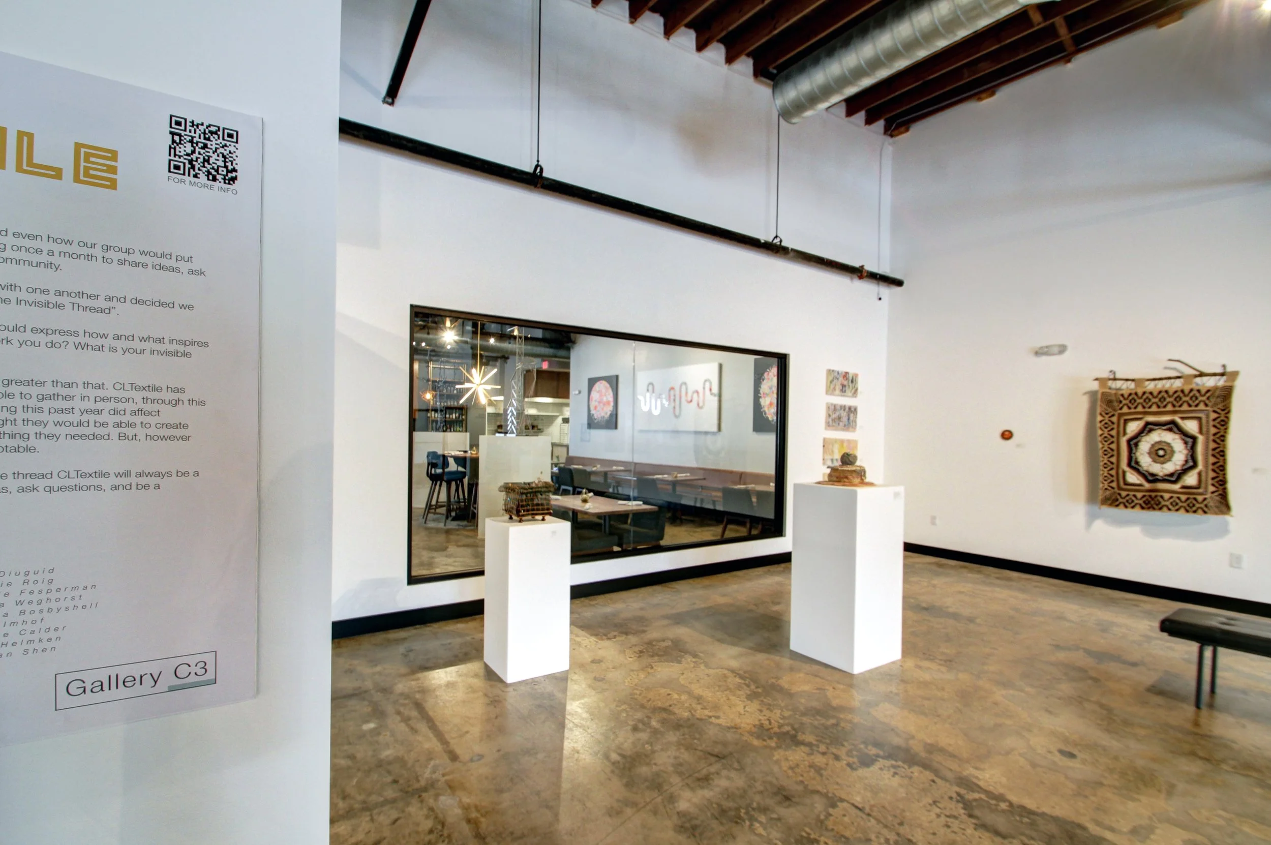 Interior of an art gallery with three sculptures on white pedestals, a wall with artwork, and a large window looking into another room with more art and seating.