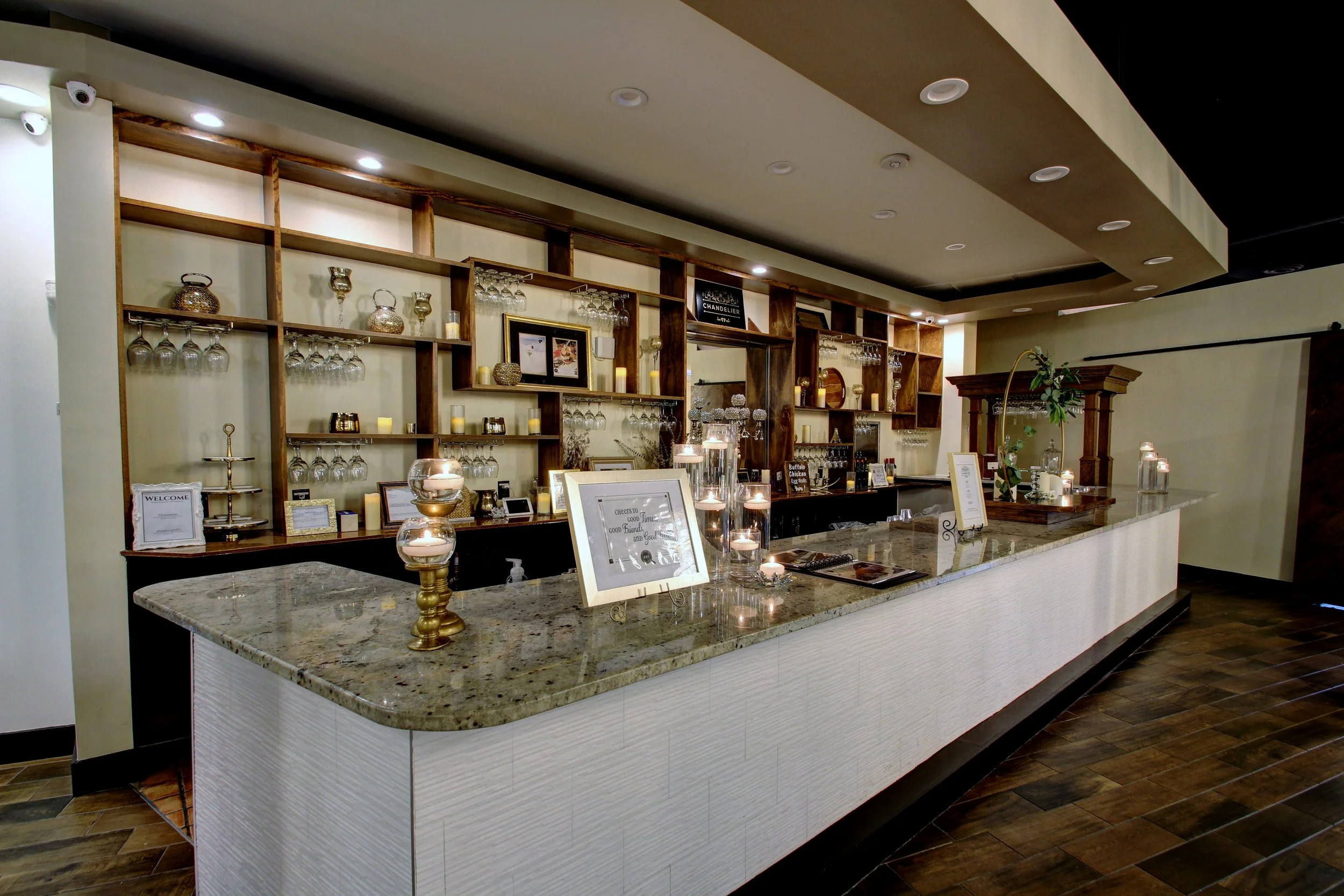 Decorative bar area with shelf displaying wine glasses, candles, and picture frames, granite countertop, and a wooden back wall with wine glass holders, in a restaurant or hotel lobby.