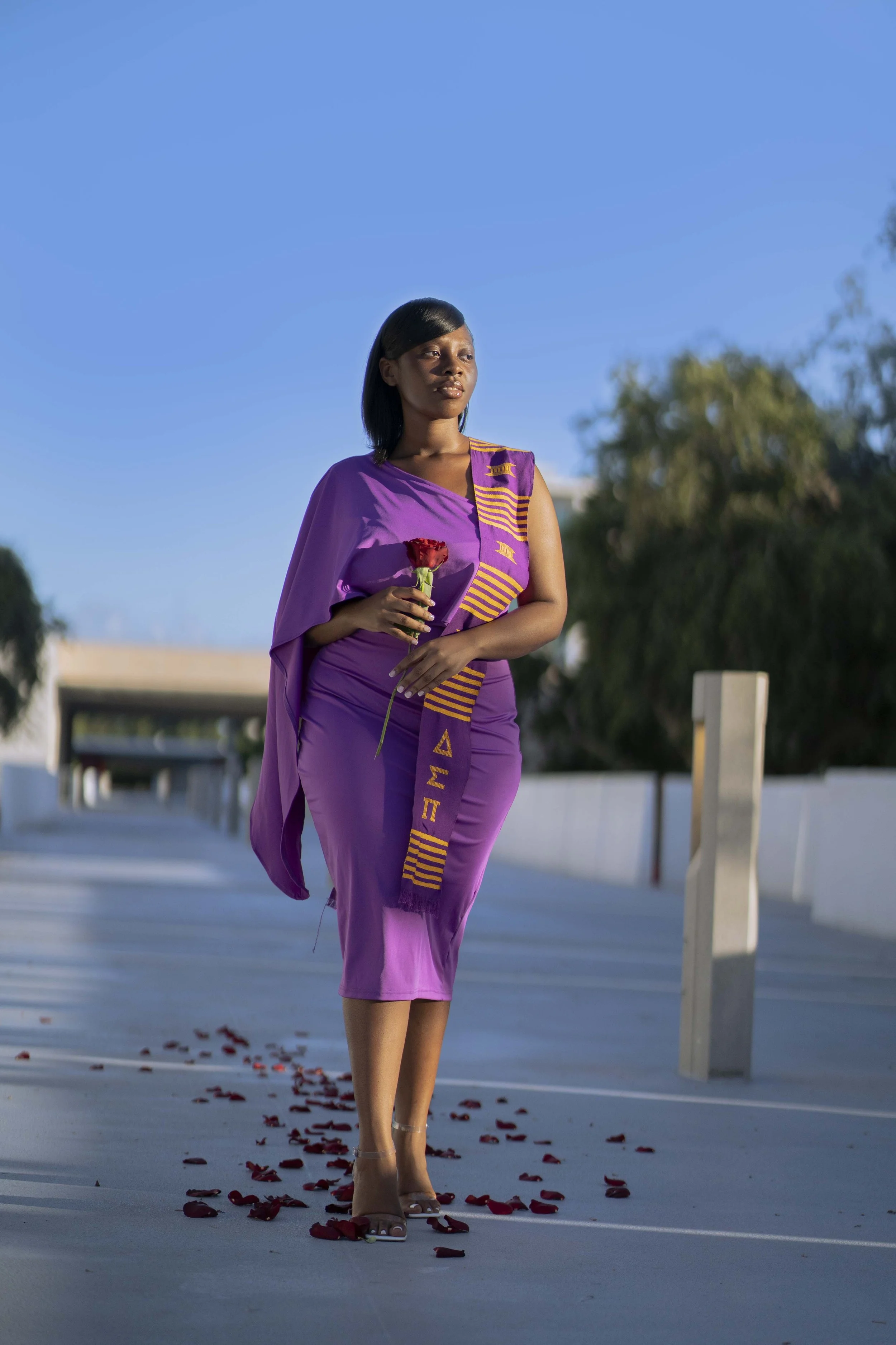 A woman wearing a purple dress and sash, holding a red rose, walking on a pathway decorated with rose petals during daytime.