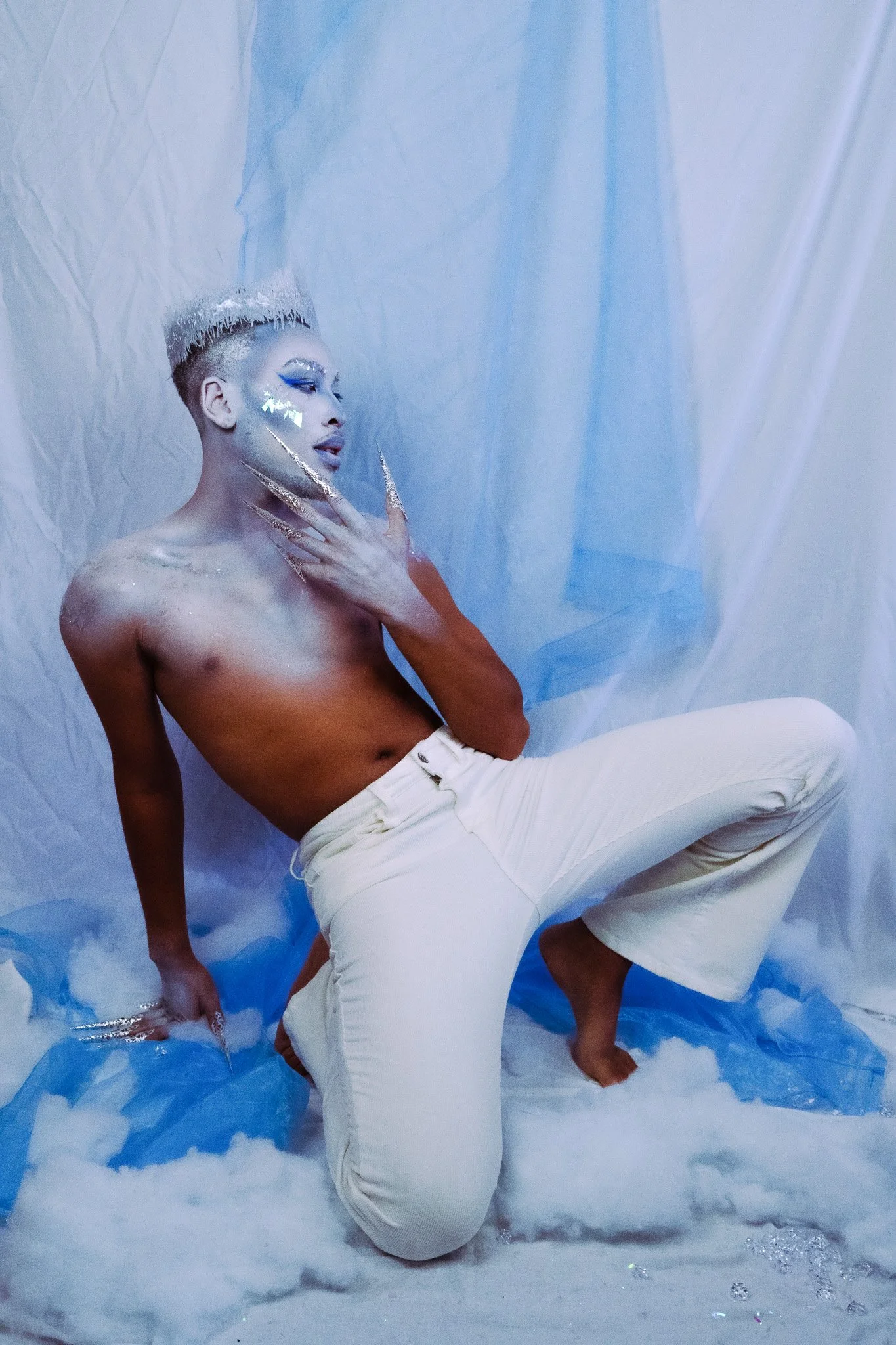 Person with artistic makeup and glitter on face, posed in a studio with blue and white fabric and clouds, wearing white pants and no shirt.