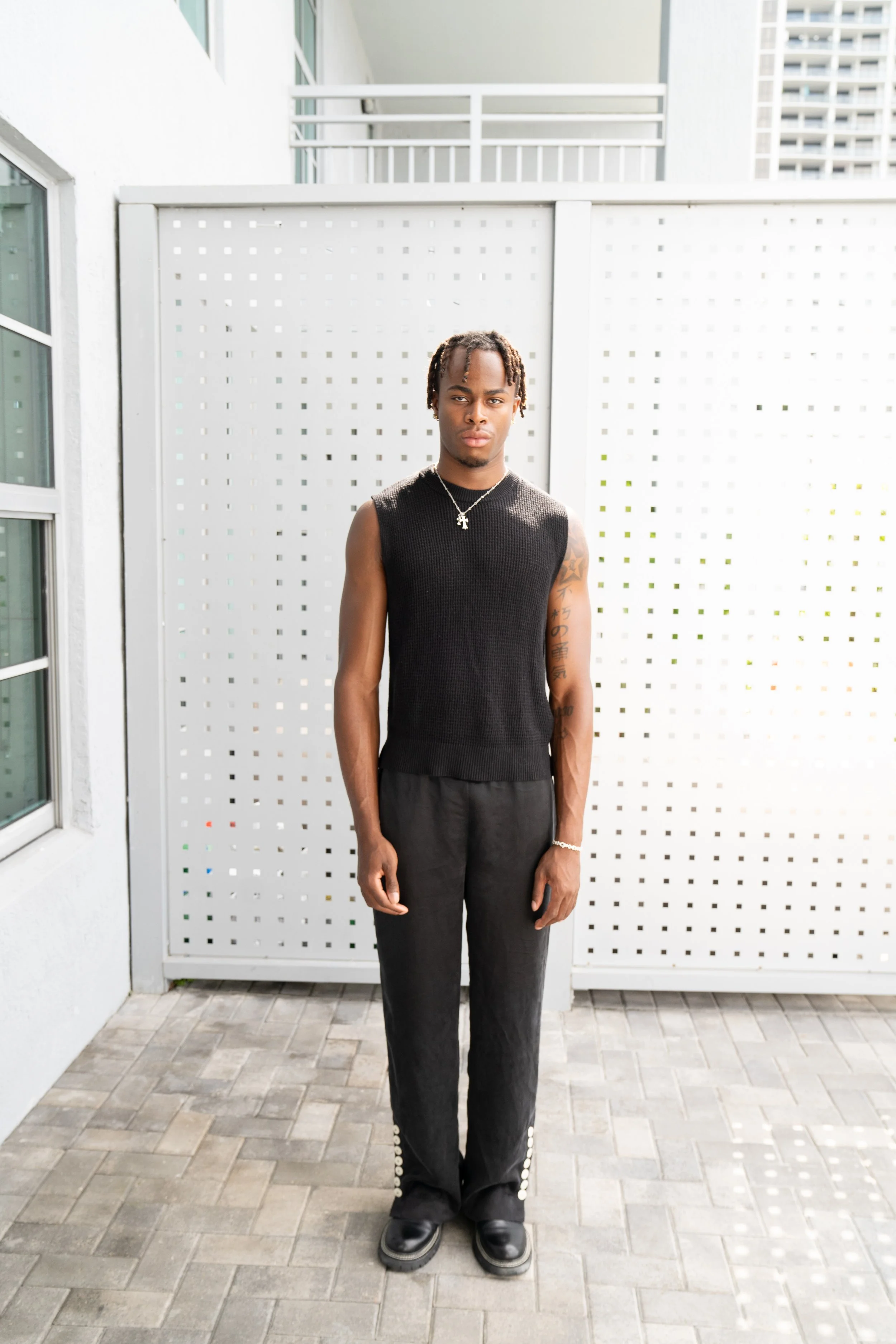 A young man in black sleeveless shirt and black pants standing outdoors against a white perforated fence.