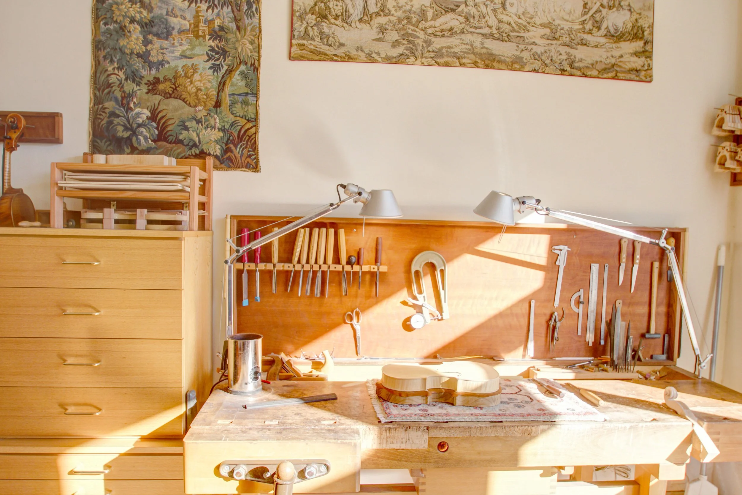 A woodworking workshop with a workbench, tools, and a partially assembled guitar. The workbench has a wooden guitar body, and various woodworking tools like chisels, scissors, and a saw hang on the wall. Sunlight shines onto the workspace.