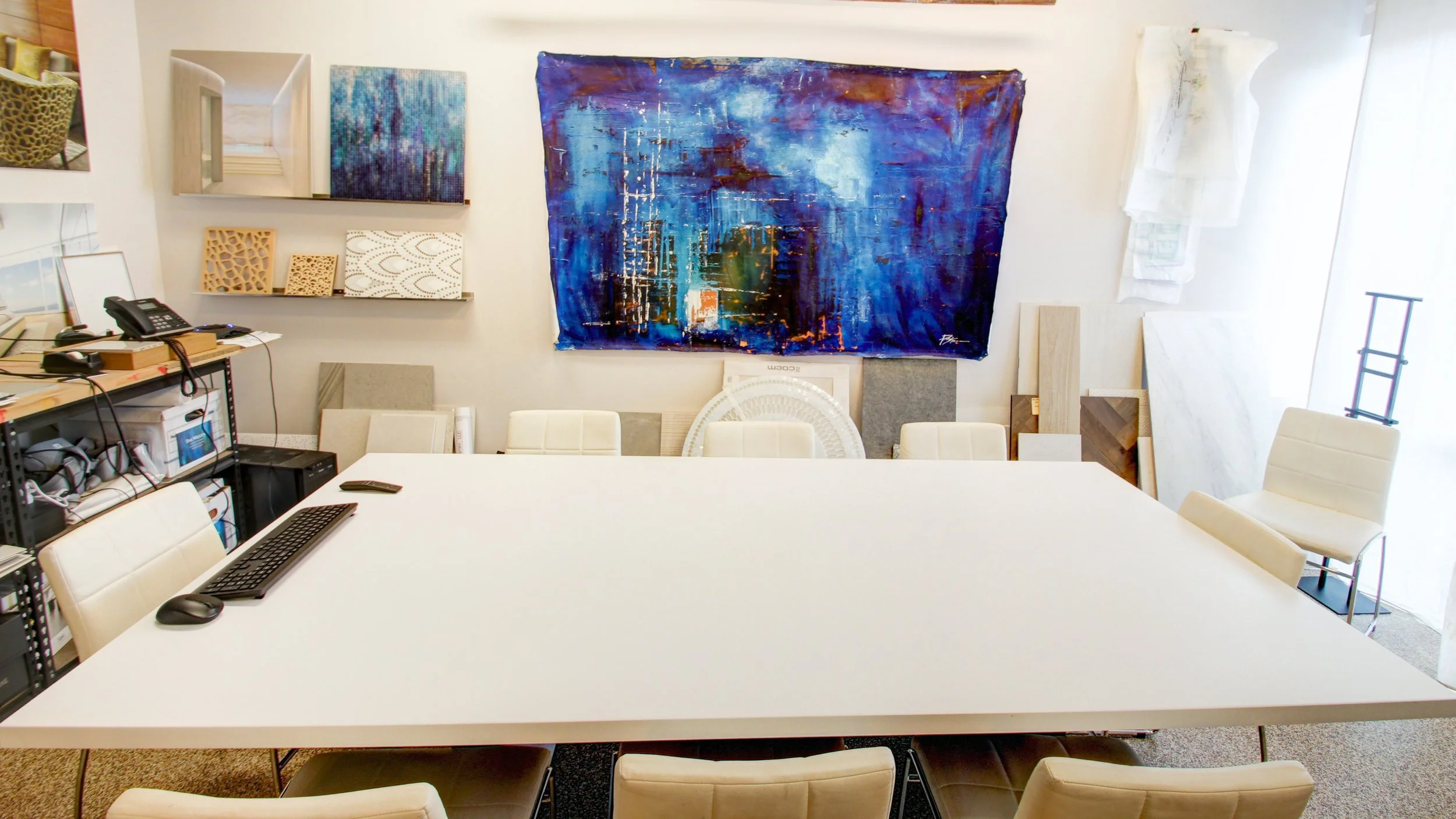 Empty conference room with a white table, white chairs, abstract artwork on the wall, and various samples of materials and wood pieces leaning against the wall.