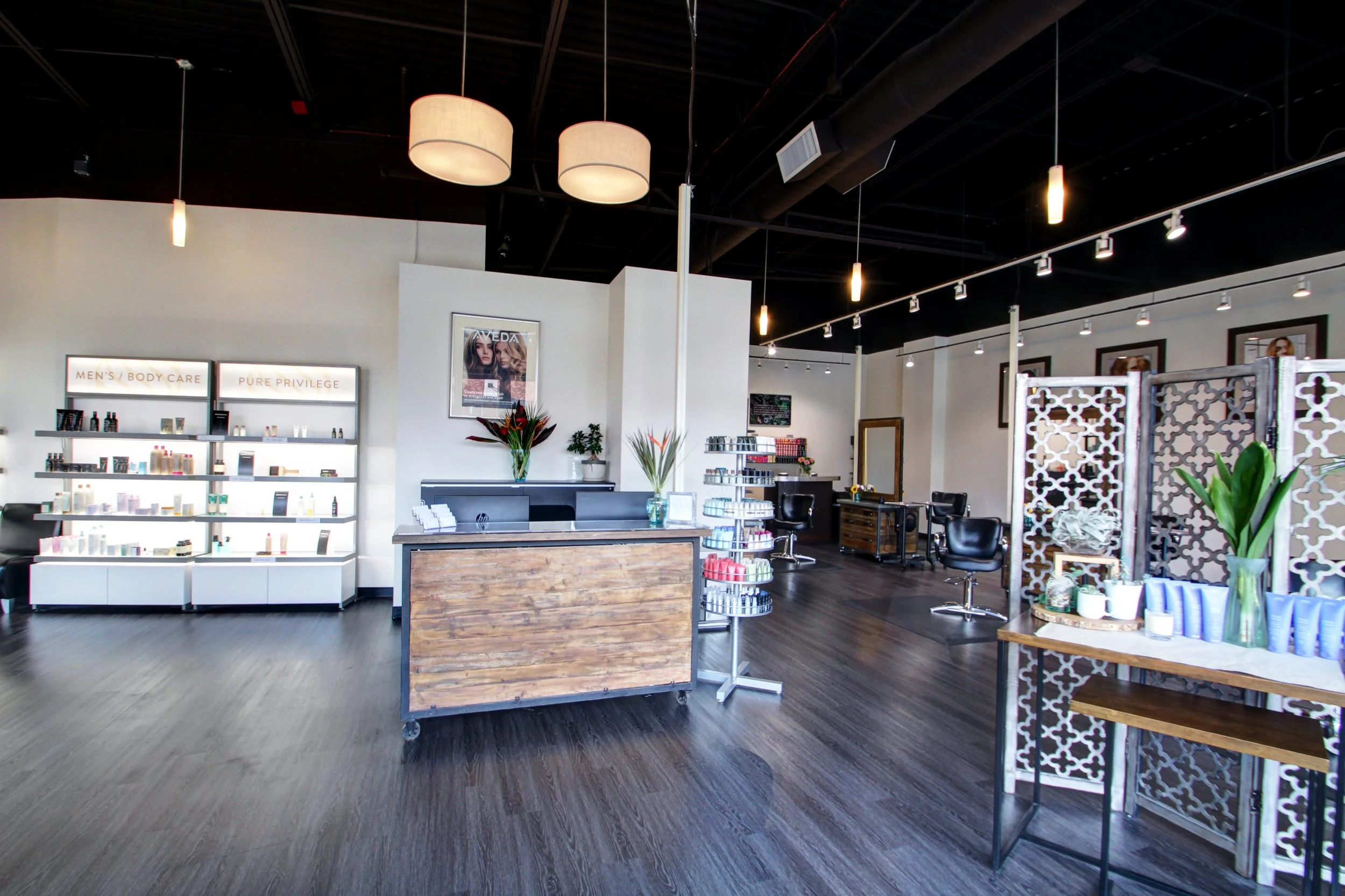 A modern salon interior with a reception desk, product shelves, styling stations with chairs, and decorative screens. Hardwood floors and pendant lighting are visible.