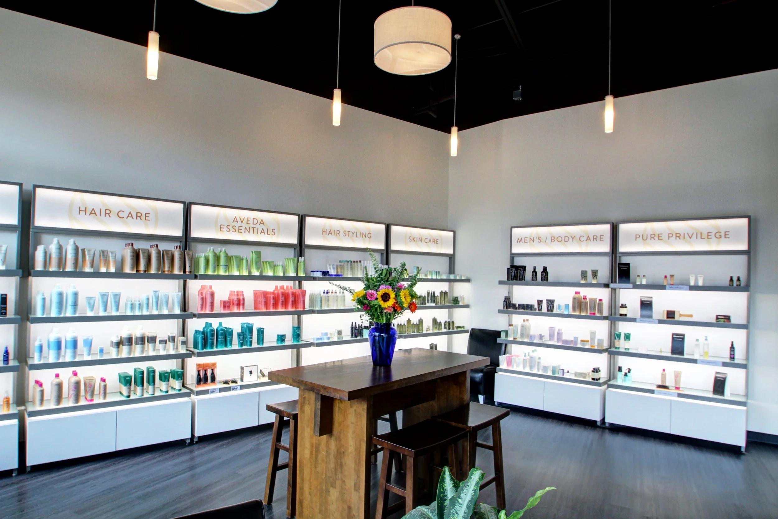 Interior of a retail store with shelves displaying hair and skin care products, a wooden table with a vase of colorful flowers, and lighting fixtures hanging from the ceiling.
