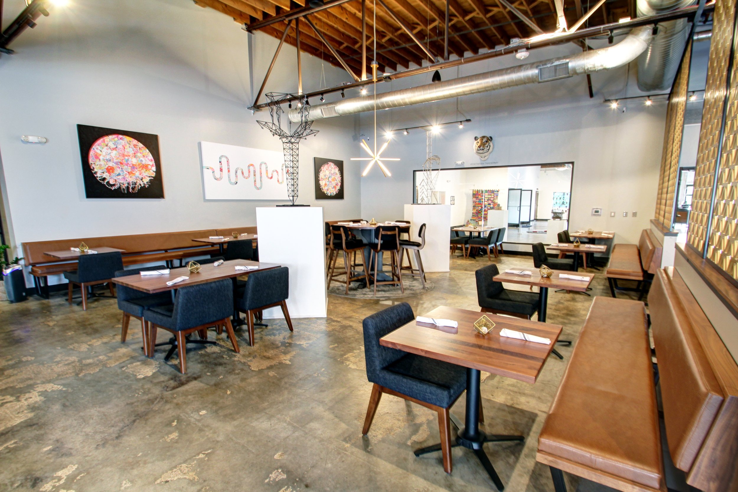 Interior of a modern restaurant with wooden tables and black and brown seating, artwork on the walls, and decorative industrial-style light fixtures.