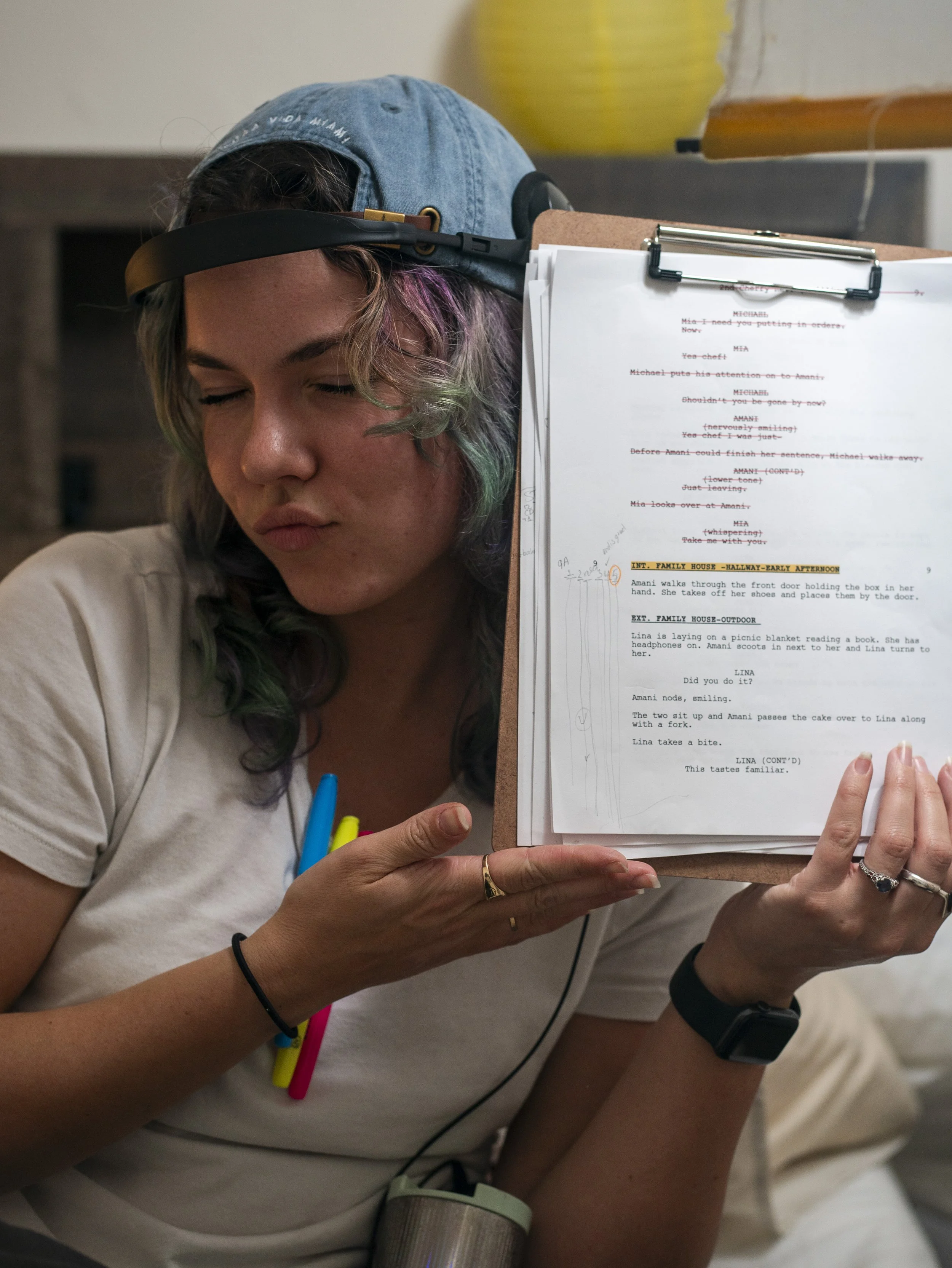 A person with multicolored hair, a denim cap, and multiple bracelets holding a script on a clipboard.