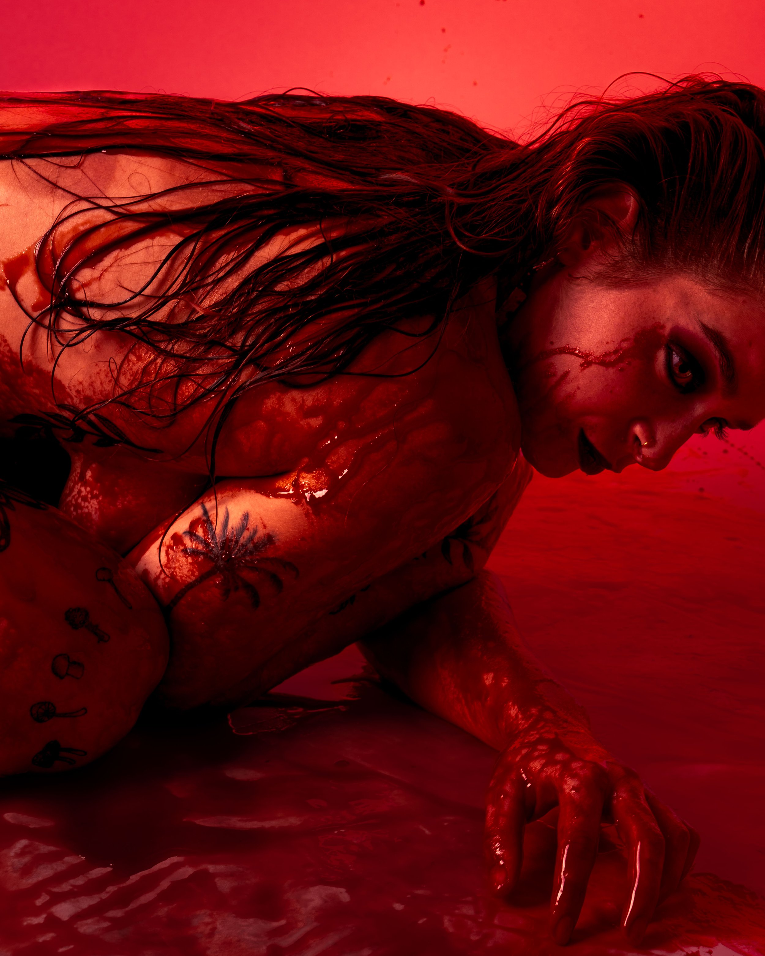 A woman with long wet hair in a red-tinted room, on her side with one arm on the ground and a menacing expression, with blood and scratches on her face and body.