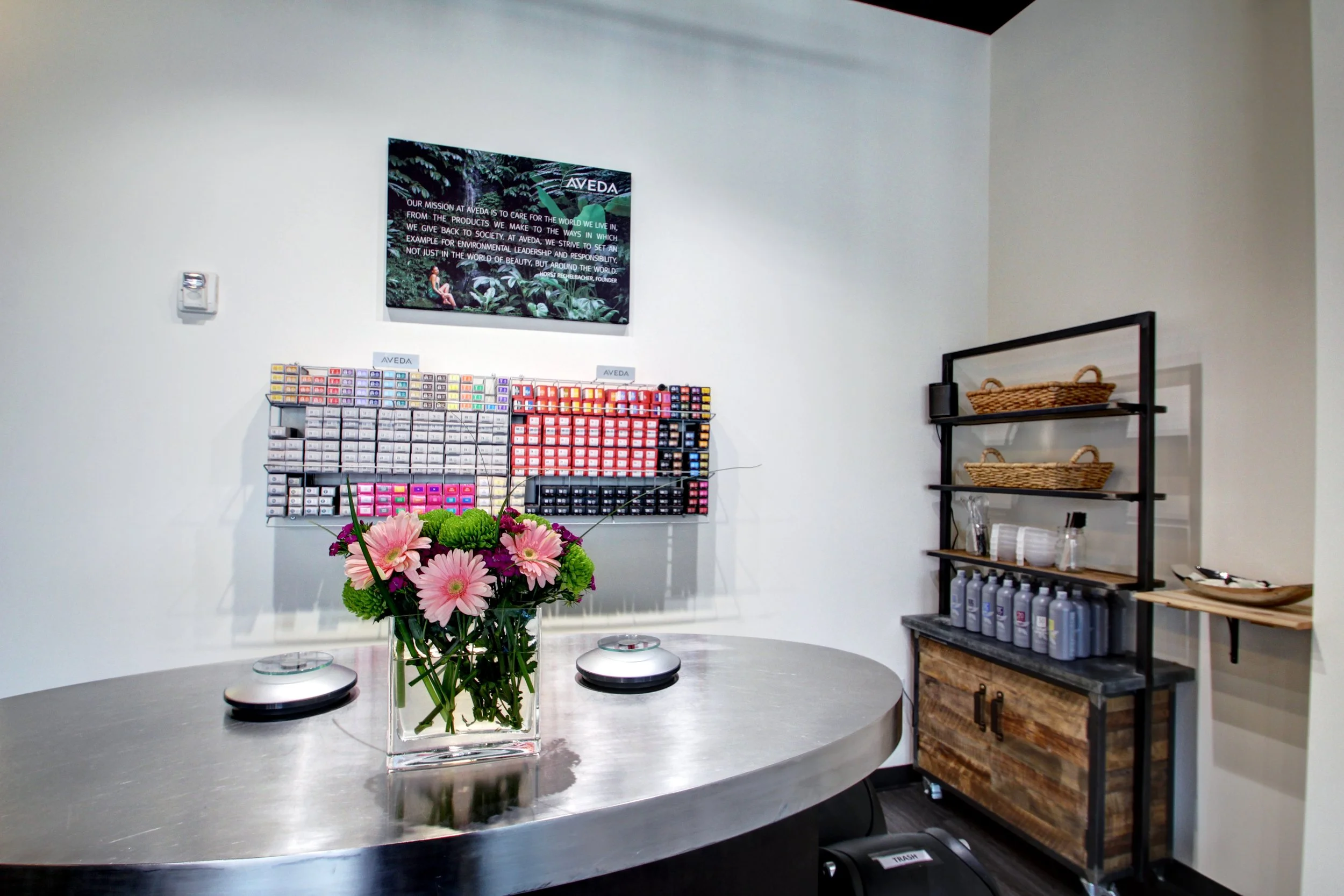 Interior of a skincare or beauty store with a round metallic table, pink flowers in a glass vase, shelving with skincare products, and a wall-mounted display of colorful product samples.