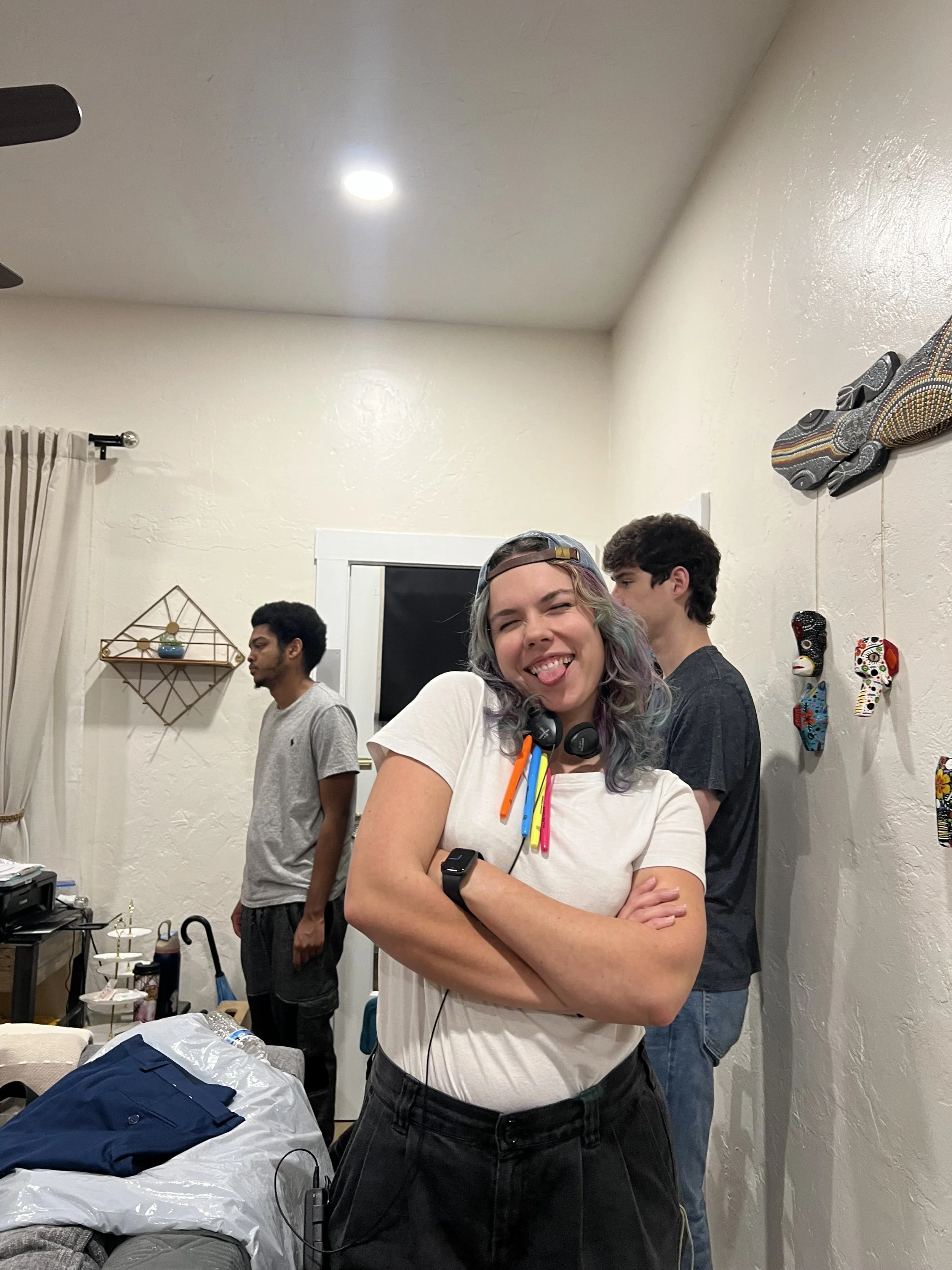 A group of young people in a room, with one smiling person in the foreground wearing a white shirt, colorful pens on their chest, headphones around their neck, and a smartwatch on their arm. The background shows three other people standing, some mask