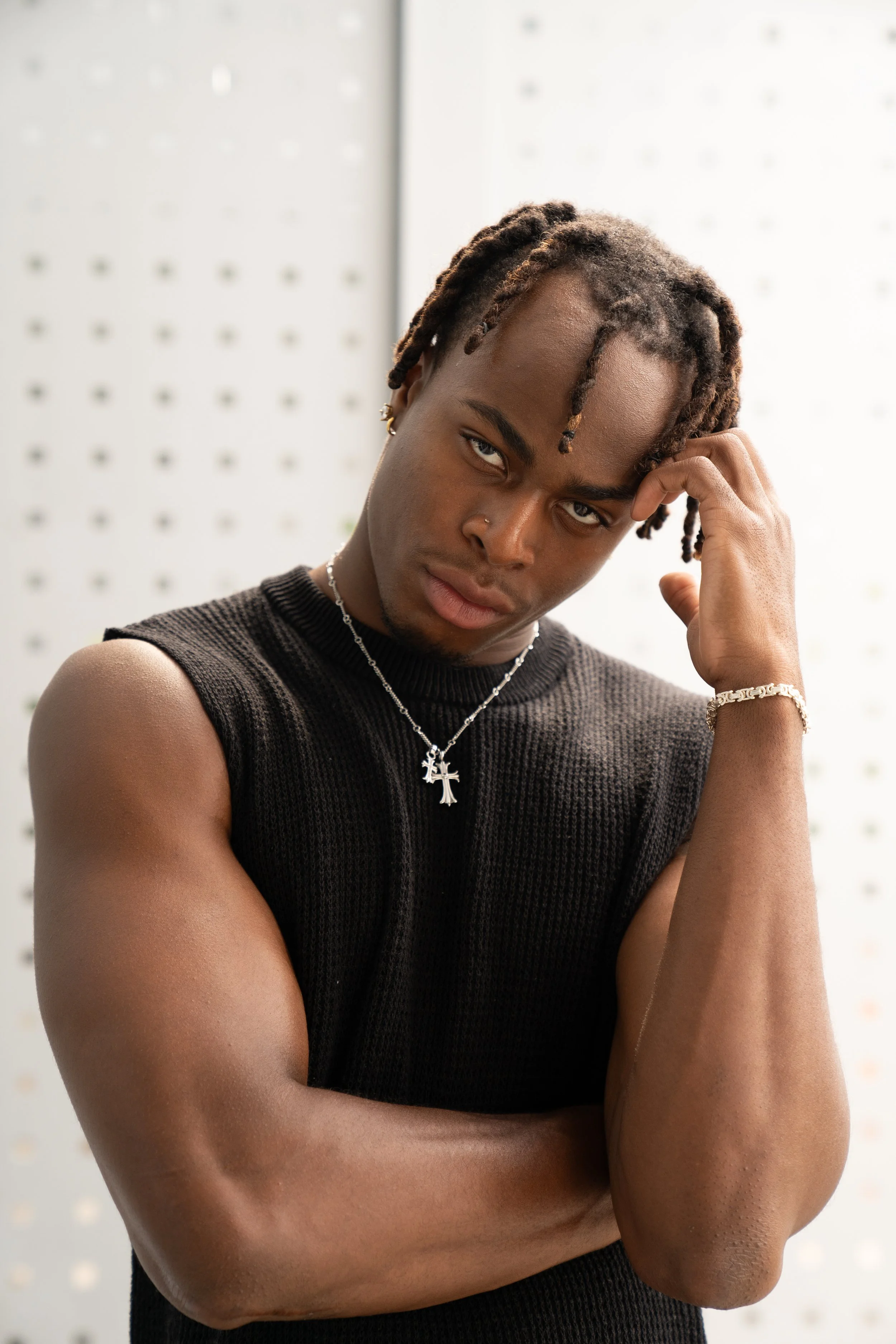 Young man with dreadlocks, wearing a black sleeveless sweater, jewelry, and a serious expression, standing in front of a white patterned background.