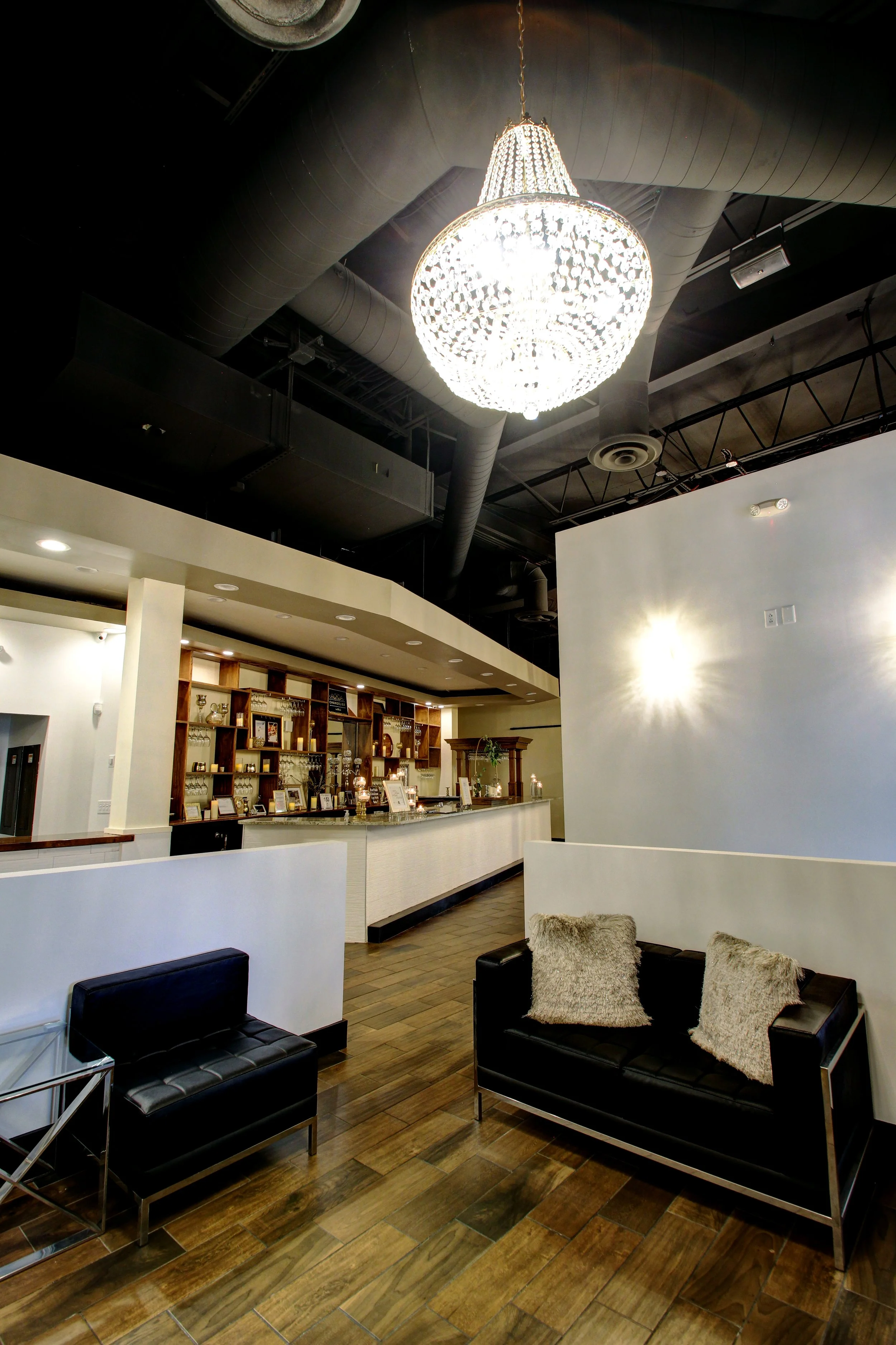 Interior of a modern bar or lounge area with a large chandelier, wooden flooring, black leather furniture, and a bar counter with shelves behind it.