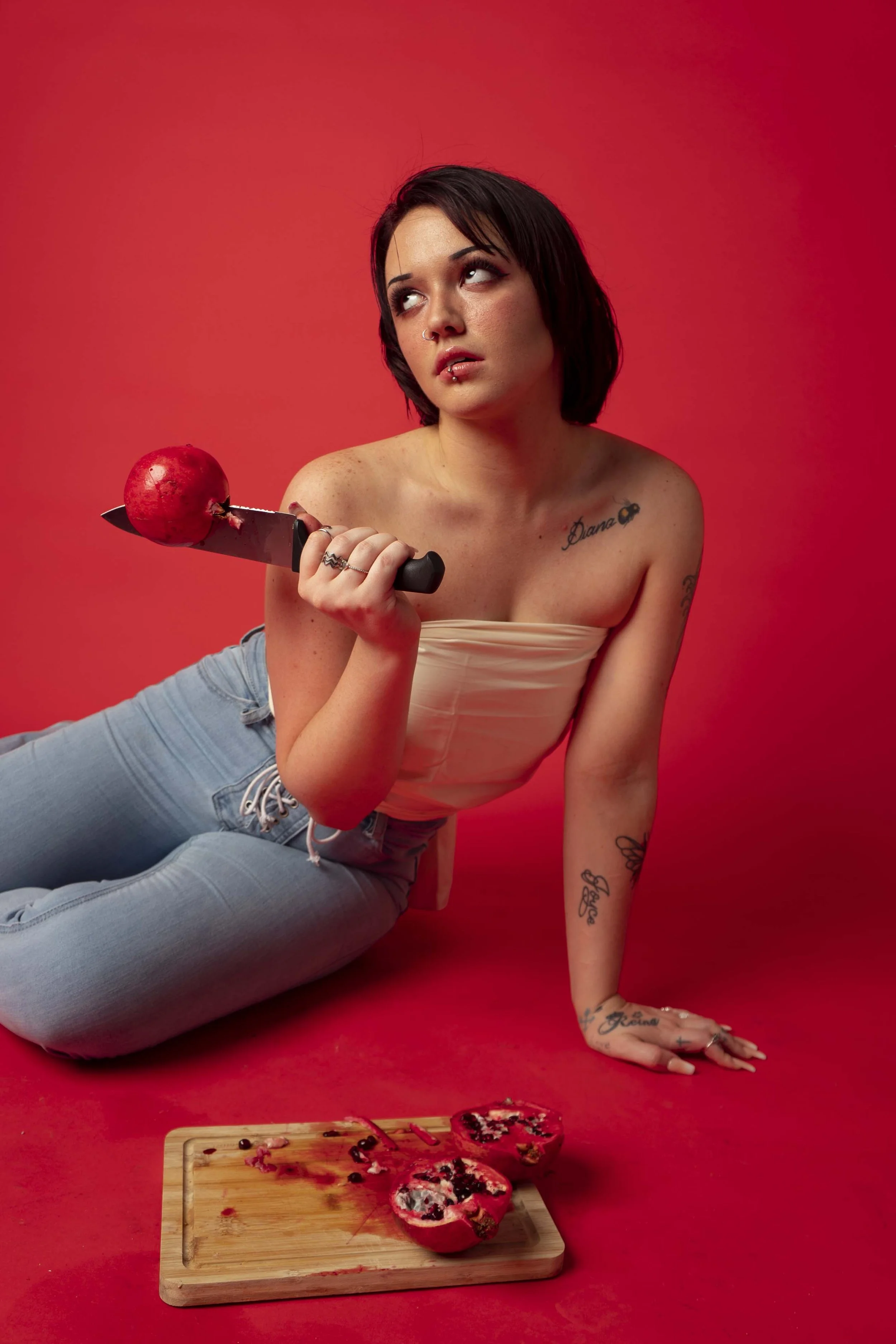 A woman with dark hair and tattoos, sitting on a red background, holding a knife with an apple on it. She has a contemplative expression, wearing a strapless cream top and jeans. There is a wooden cutting board with a halved pomegranate in front of h
