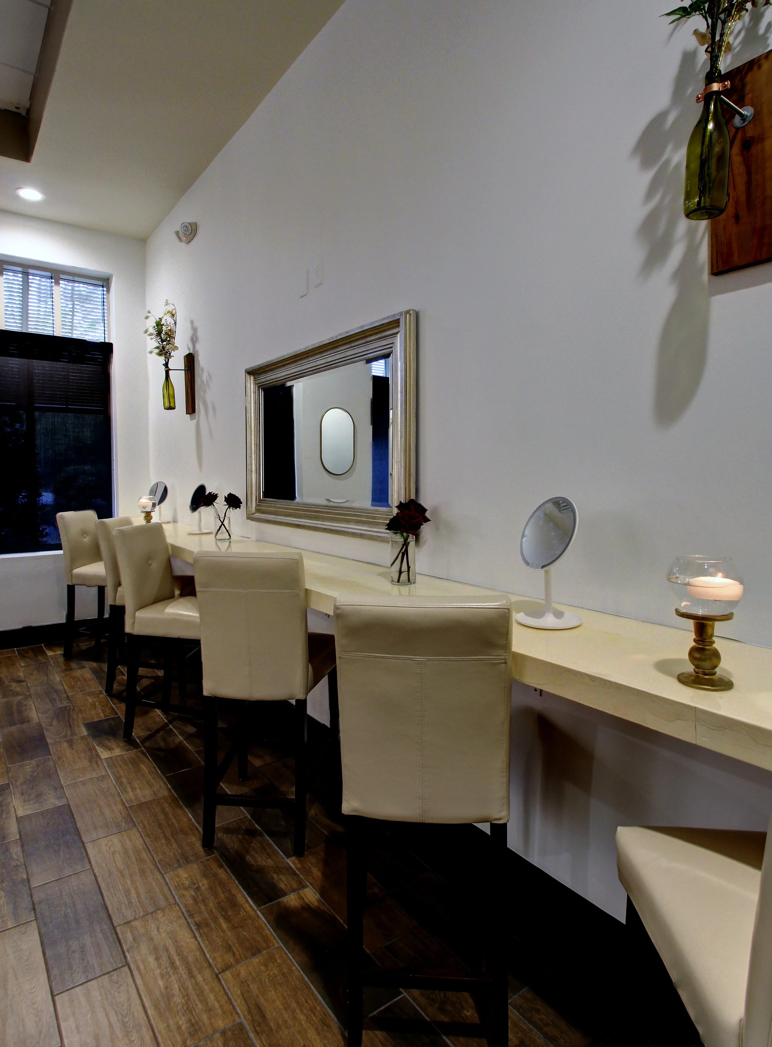 Cream-colored bar stools lined up against a long cream-colored counter with decorative vases and small mirrors on top, a large mirror on the wall, and a wooden floor.