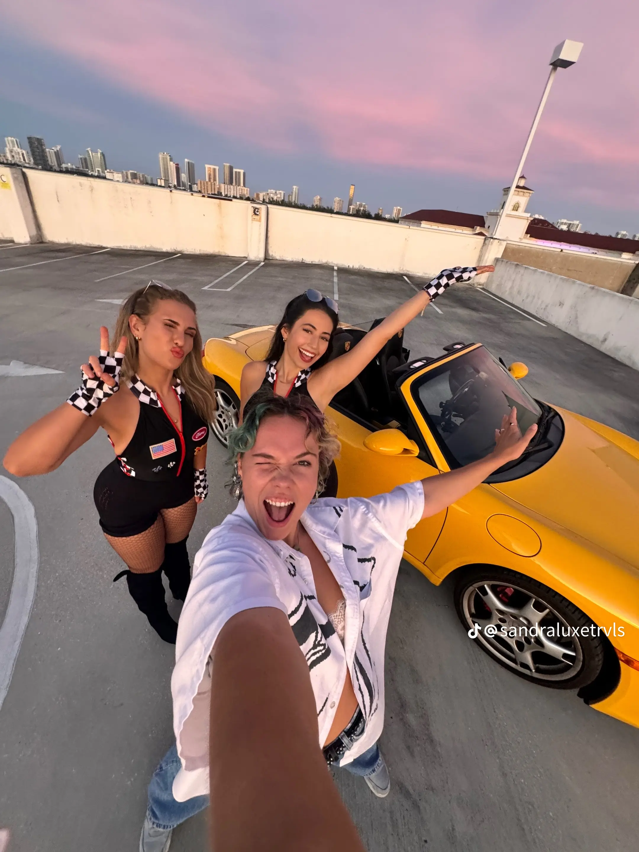 Four friends taking a selfie in a parking garage with a yellow convertible car and city skyline in the background during sunset.