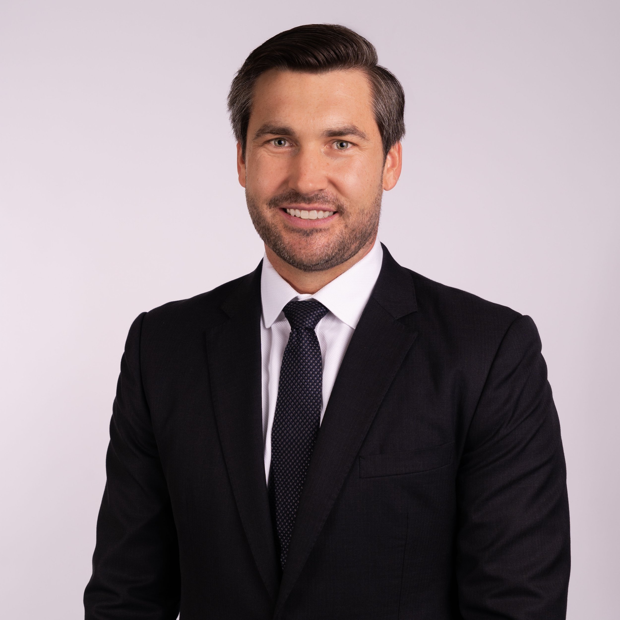 A professional man in a black suit, white shirt, and black tie, smiling and posing against a light purple background.