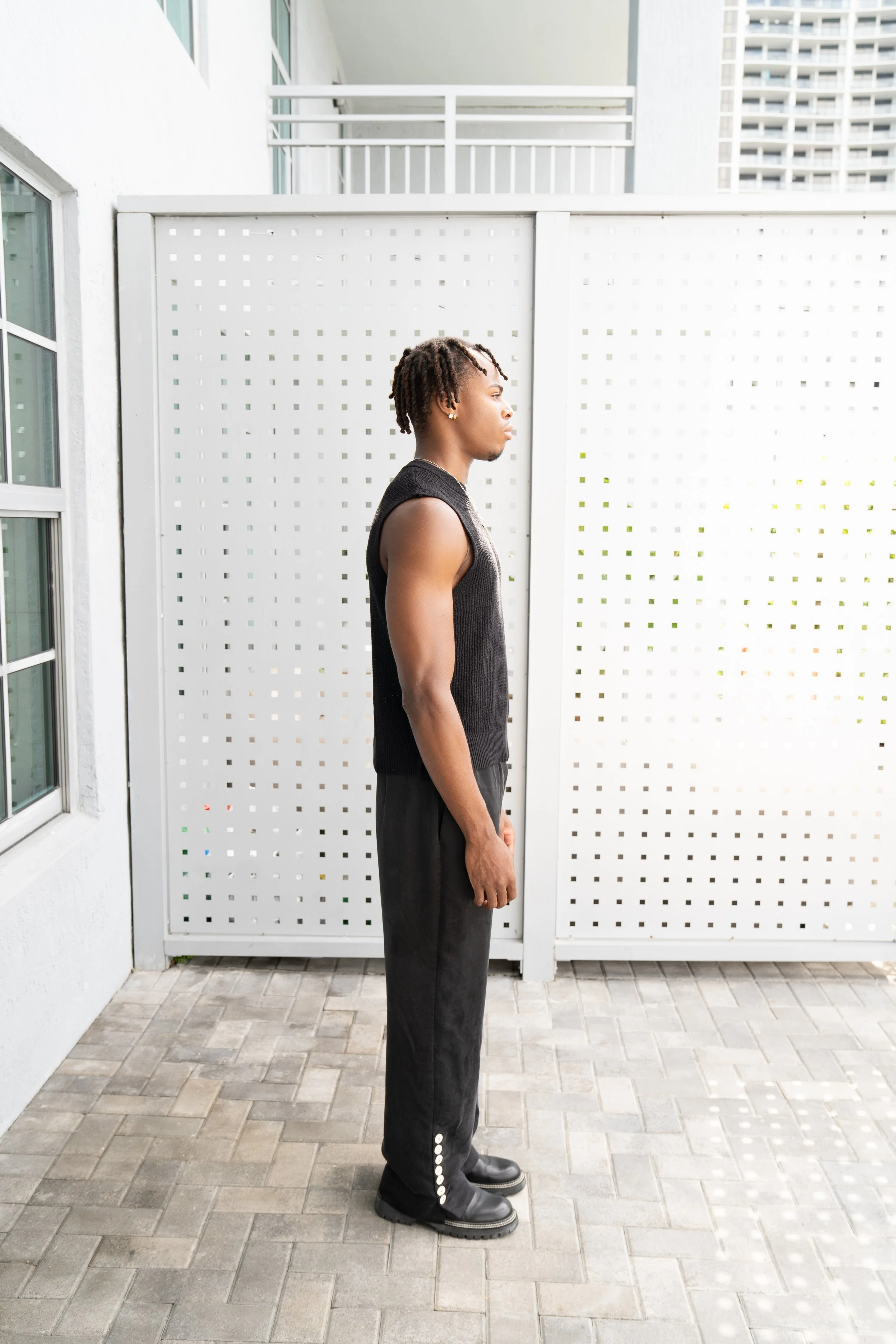 A young man with dreadlocks standing sideways outdoors against a white lattice fence, wearing a black sleeveless sweater, black pants with buttons on the side, and black boots.