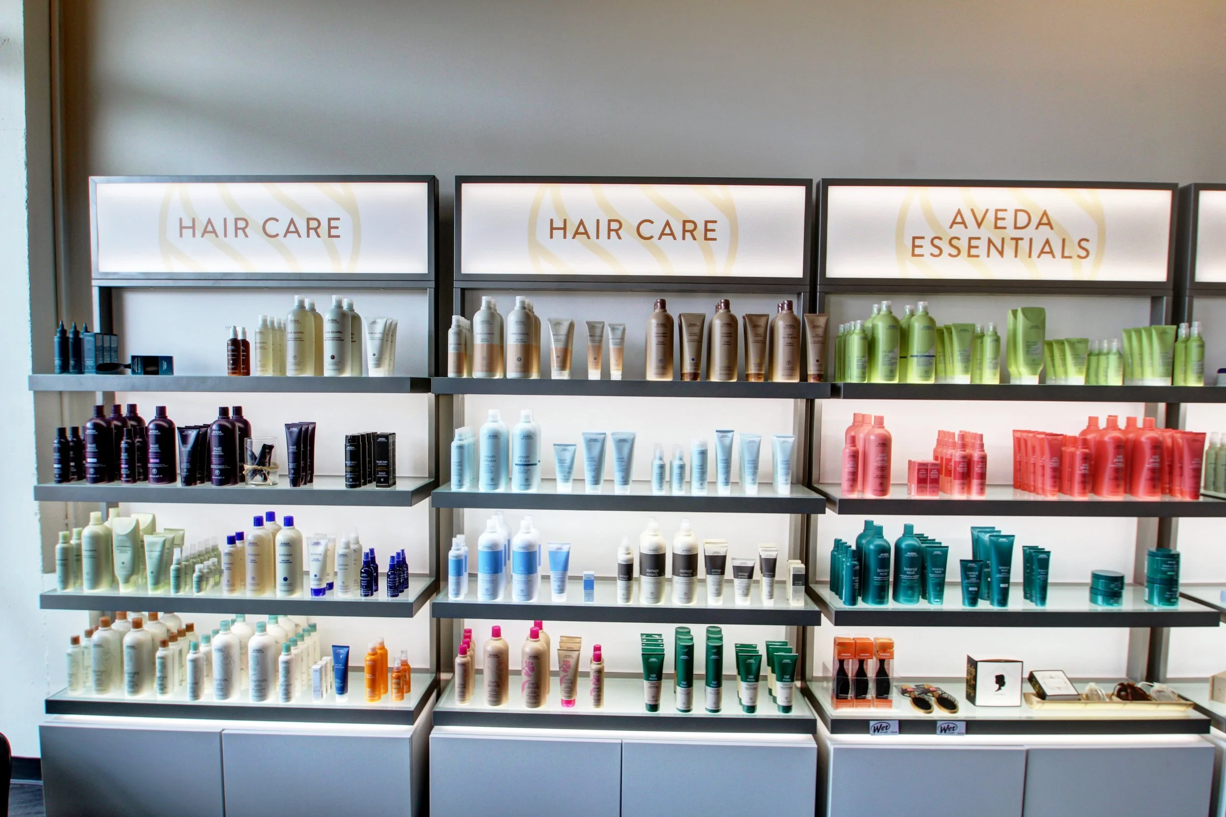 Shelves with various hair care products at a store, including shampoos, conditioners, and treatments, organized under signs labeled 'Hair Care' and 'Aveda Essentials'.