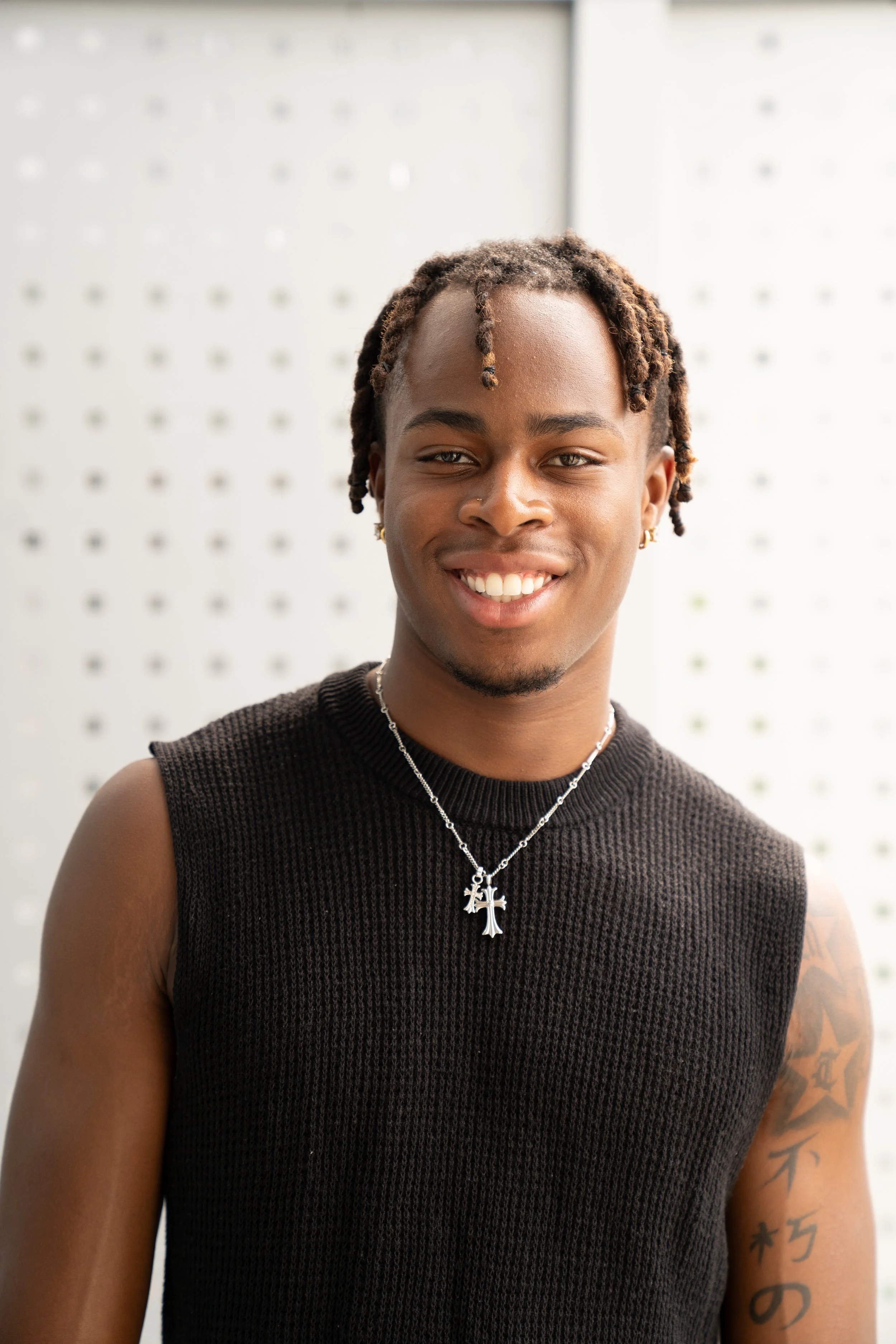 A young man with short dreadlocks, wearing a sleeveless black knit top, smiling, with a necklace featuring a cross pendant, standing in front of a perforated metal background.