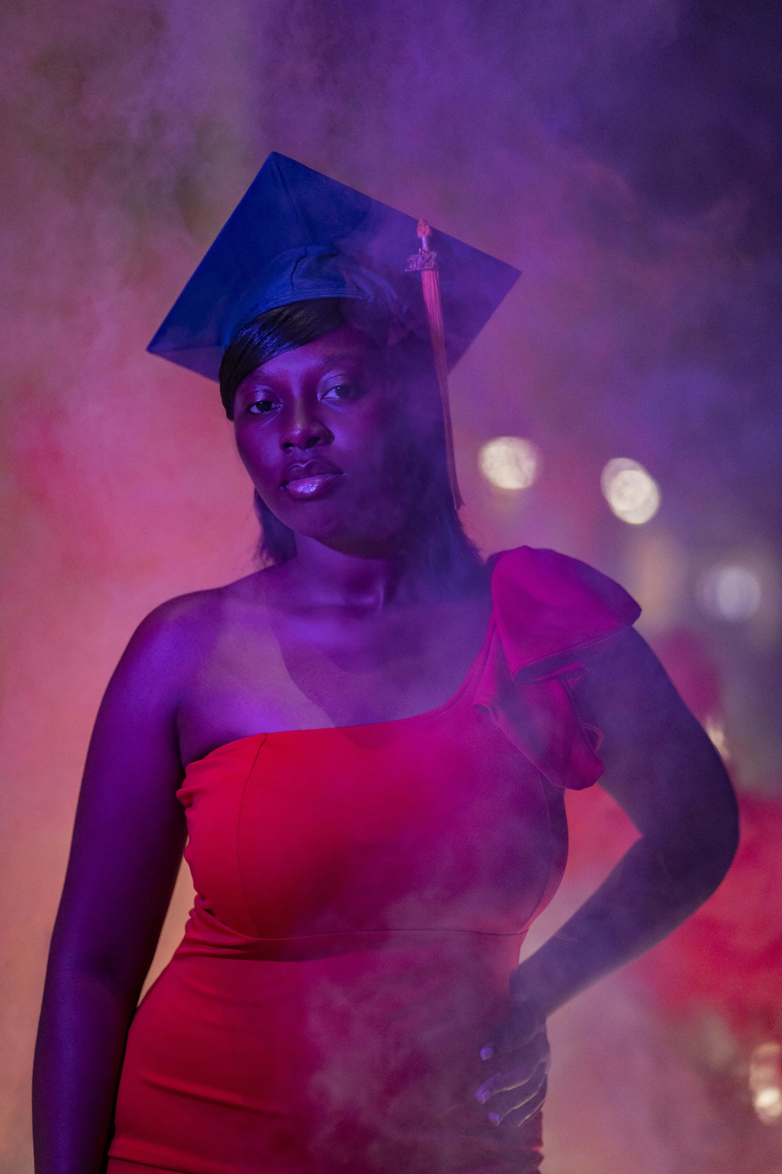 A woman in a red dress and a graduation cap stands amidst colorful smoke and lights.