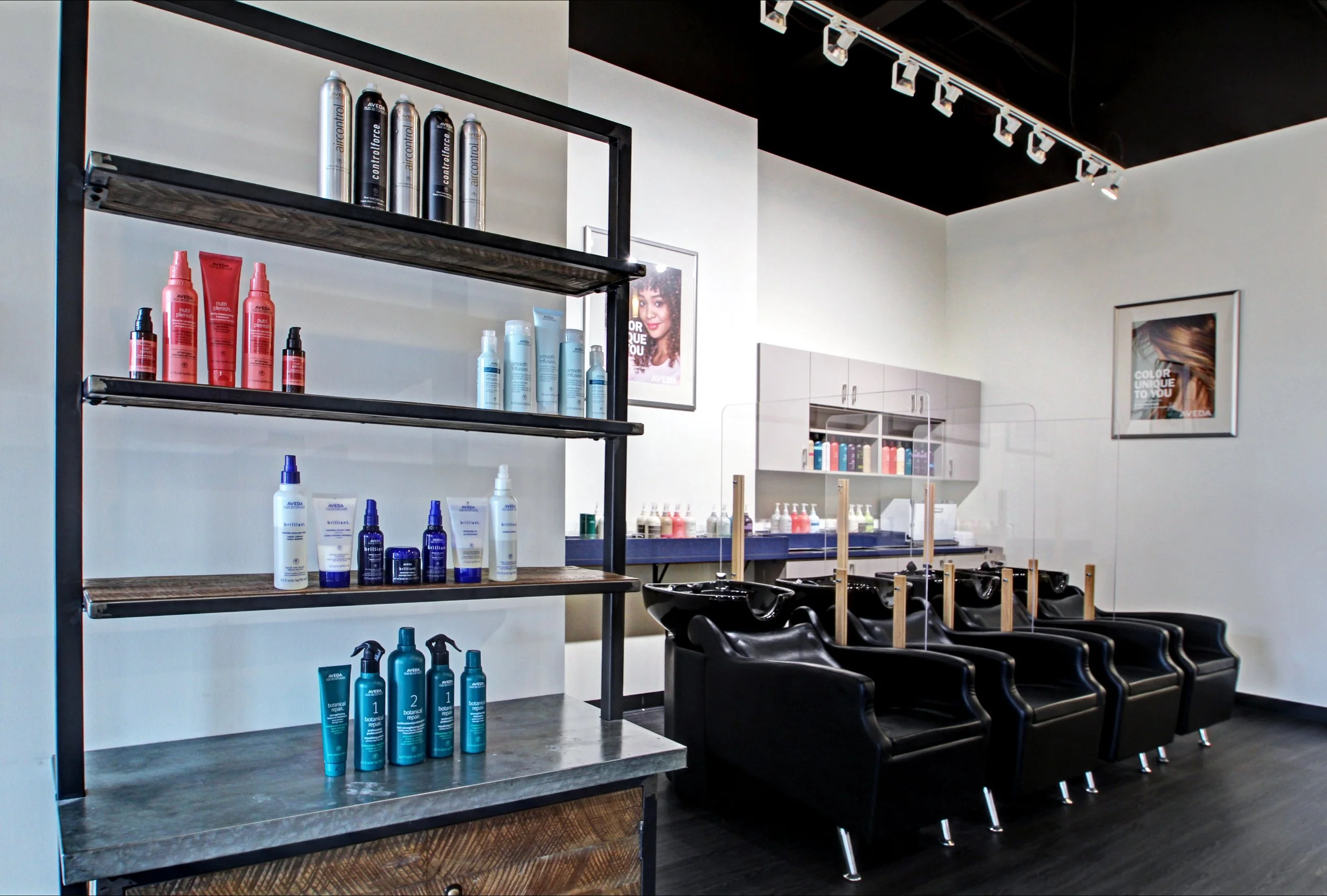 Hair salon with black chairs at washing stations, shelves with hair products, framed artwork on the walls, ceiling track lighting.