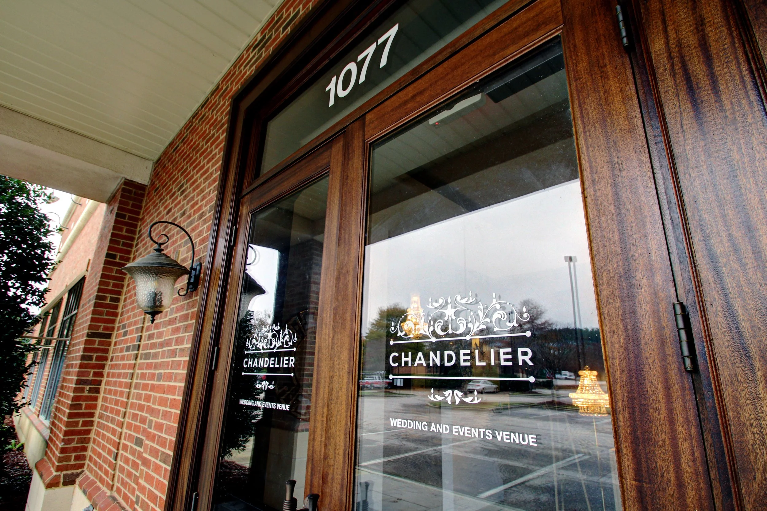 Entrance to a wedding and events venue called Chandelier, with the street number 1077 displayed above the door. The door has glass panes with the venue's logo and name, and there is a brick exterior wall with a wall-mounted lantern-style light fixtur