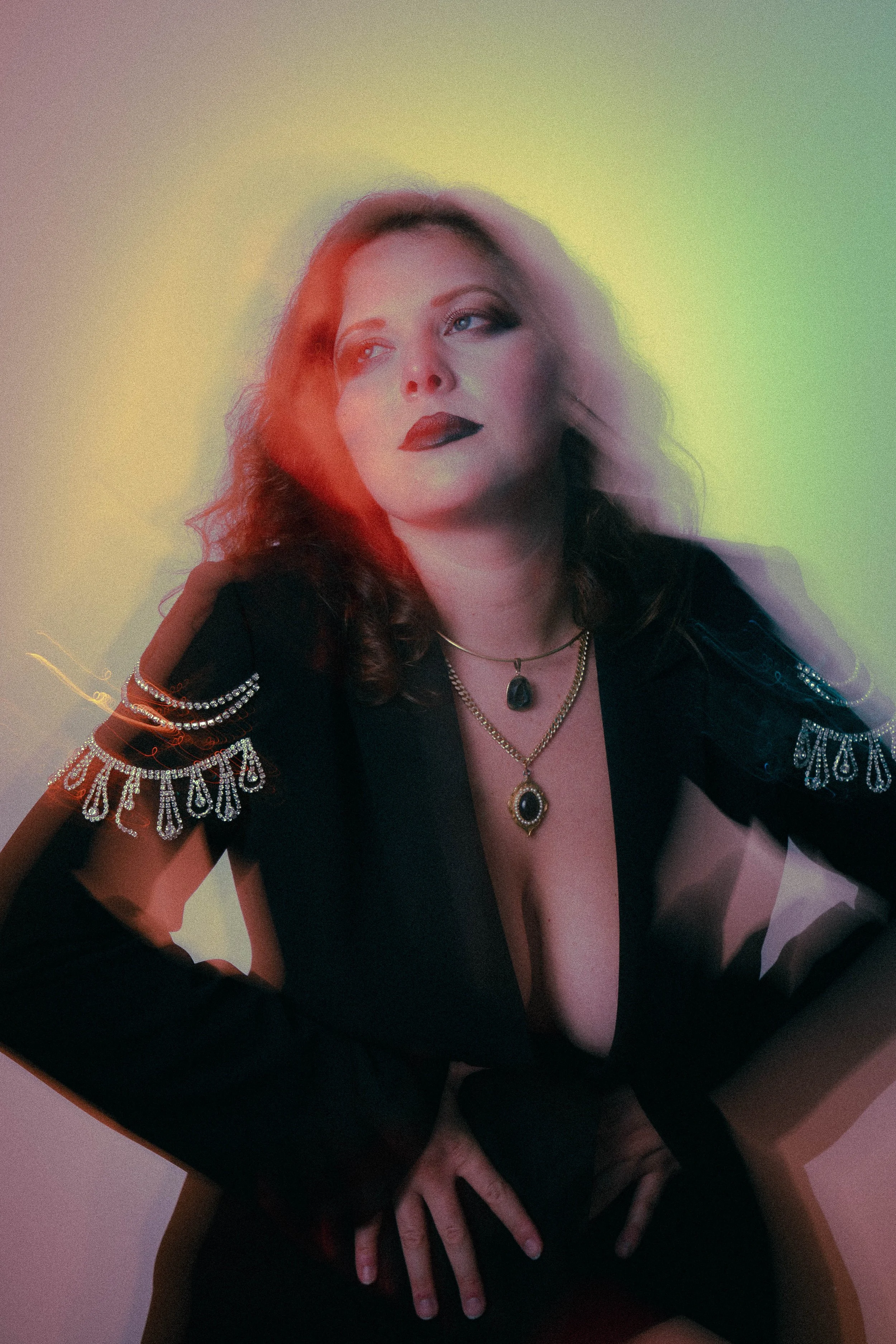 A woman with wavy hair wearing dark lipstick and layered necklaces poses with her hands on her hips against a gradient background with colorful light effects.