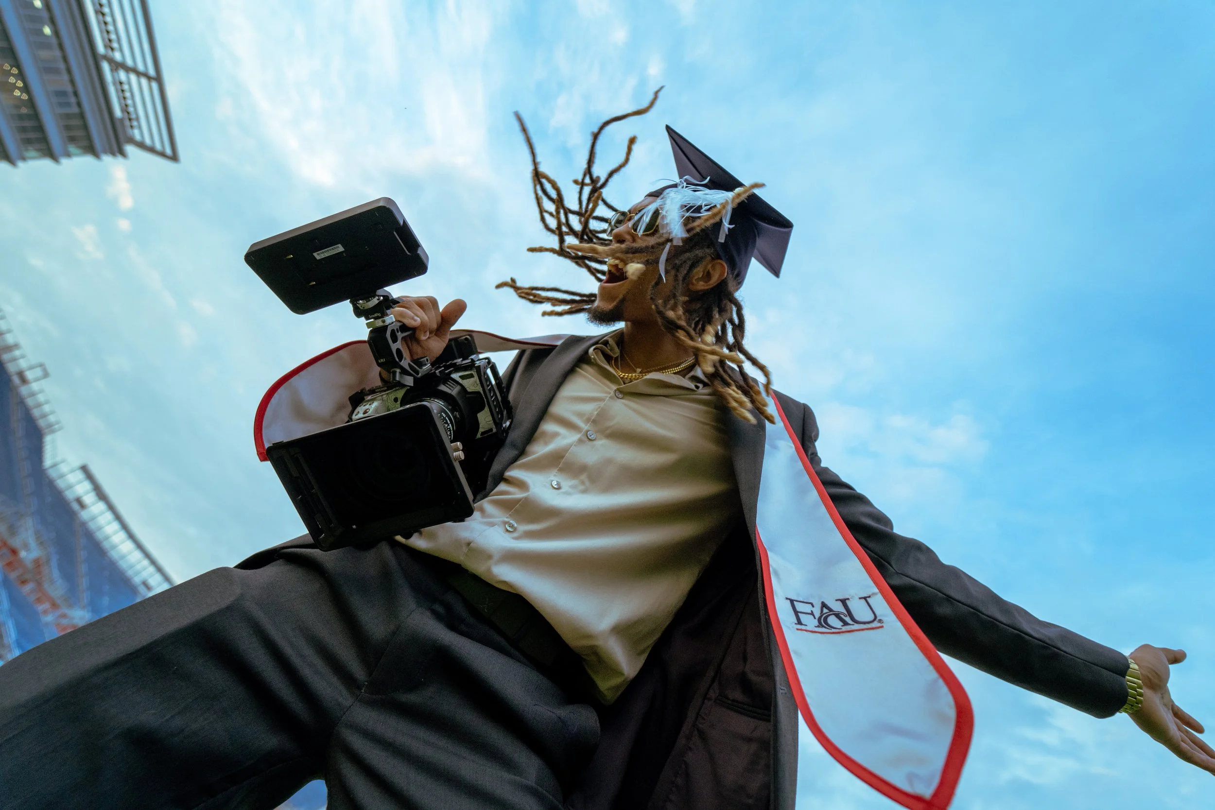 A man with dreadlocks wearing a graduation cap and gown holding a camera, smiling under a blue sky.