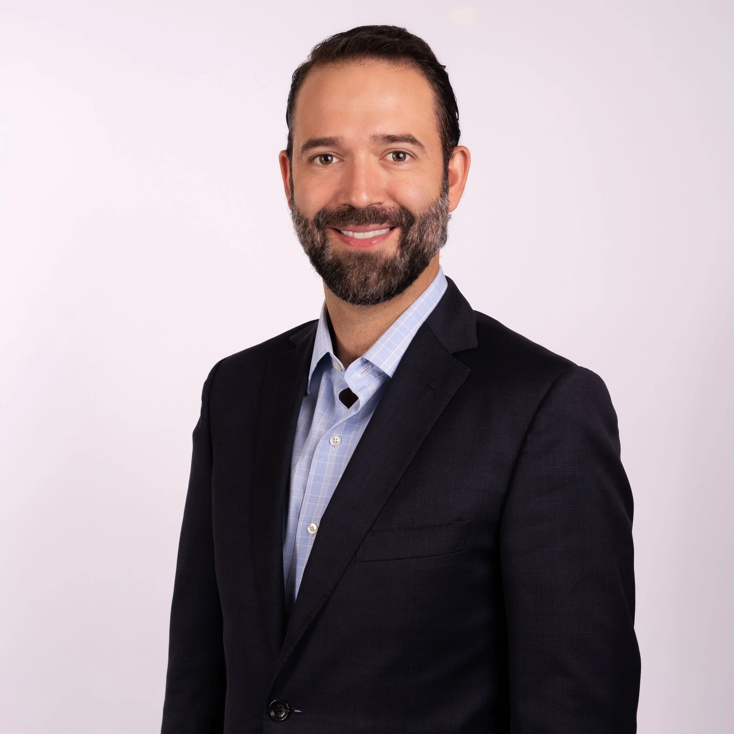 Headshot of a smiling man with a beard, dark hair, wearing a dark suit and light blue shirt against a plain light background.