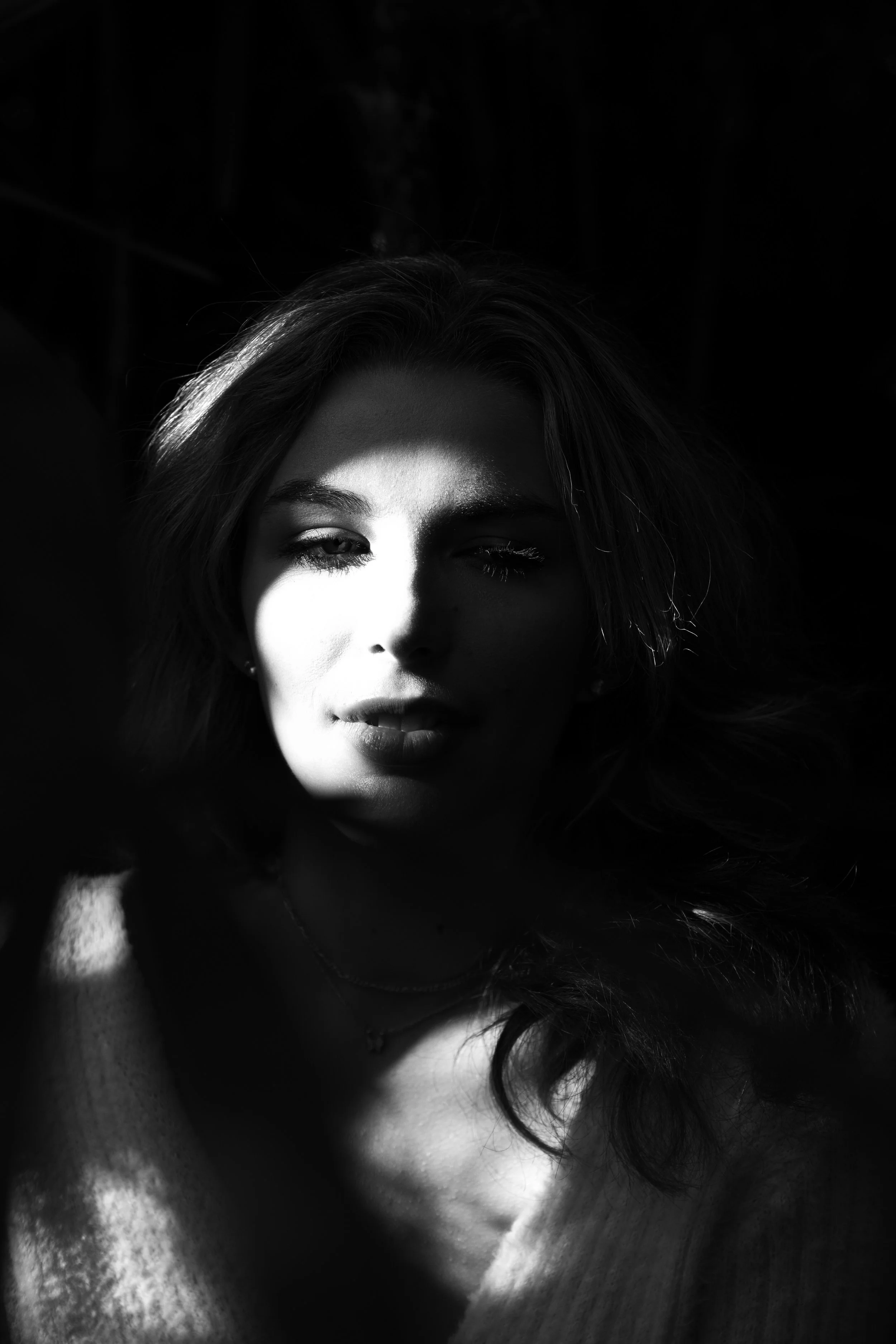 A black and white portrait of a woman with light and shadow contrasting on her face, highlighting her facial features and hair.